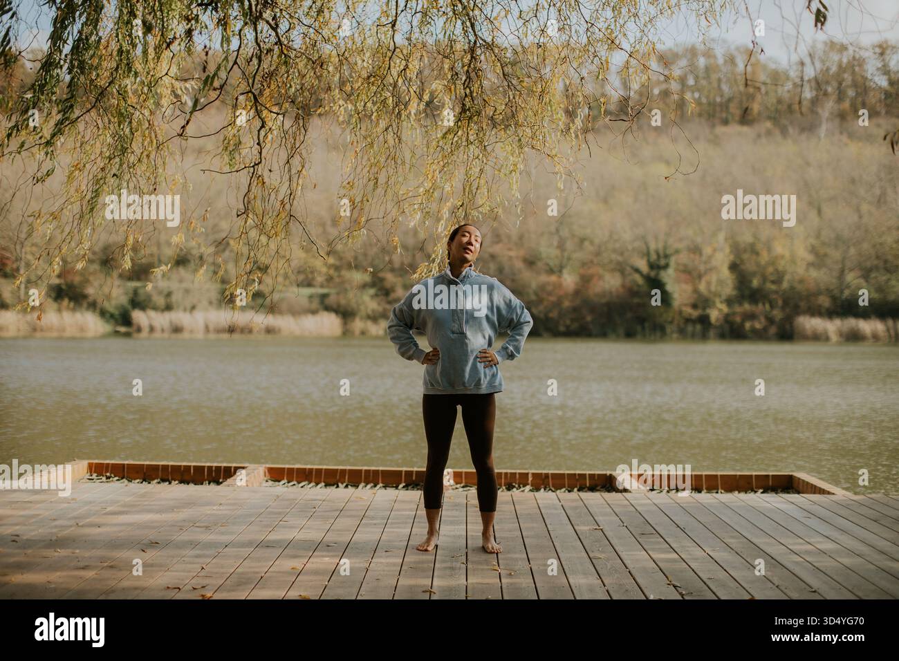 Una donna coreana si erge a piedi nudi su una piattaforma di legno vicino a un tranquillo lago circondato da alberi autunnali. Sembra rilassata e contenta. Foto Stock