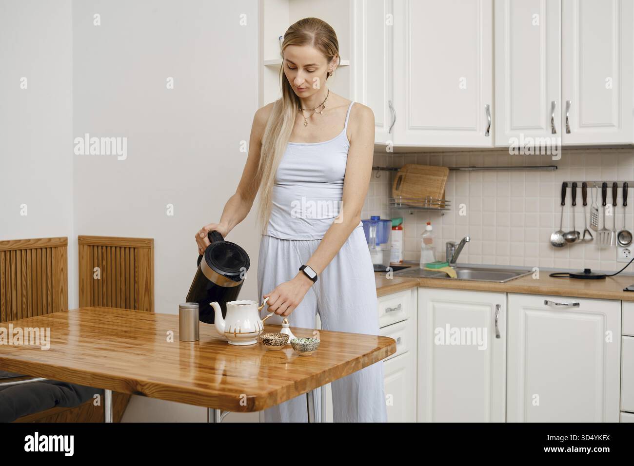 Una donna si trova in una cucina moderna che versa acqua calda da un bollitore in una teiera. Indossa un vestito leggero e si sta preparando a preparare il tè, concentrandosi su di lui Foto Stock