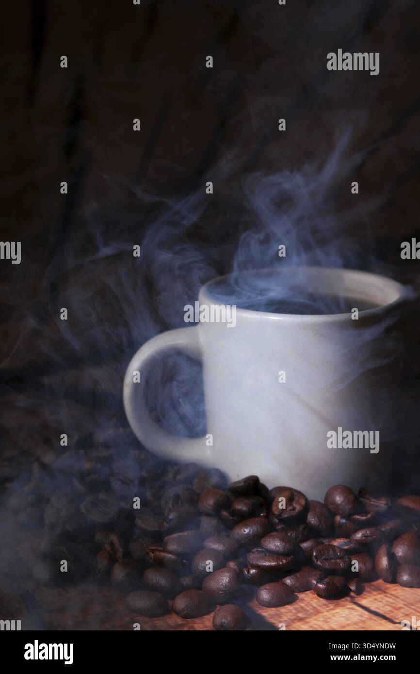 Cottura a vapore di caffè caldo in una tazza bianca in mezzo a chicchi tostati sparsi su una superficie rustica in legno scura, atmosfera calda e aromatica Foto Stock