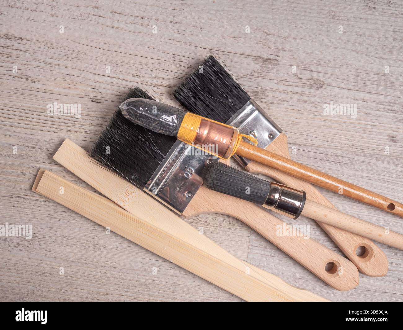 Un gruppo di pennelli assortiti con manico in legno e bastoncini di agitazione si trova su una superficie a grana di legno chiaro. Foto Stock