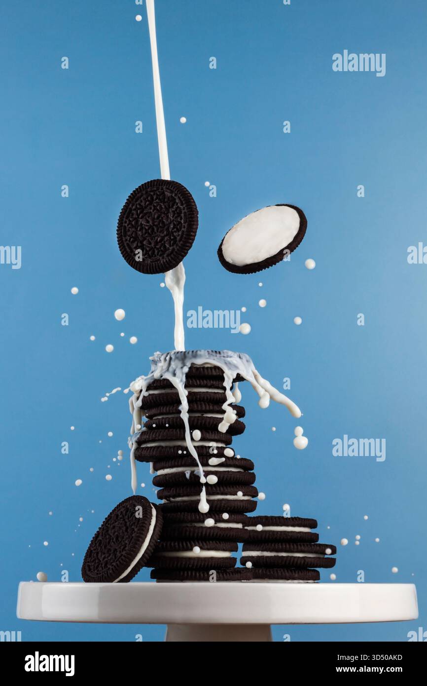 I biscotti sbriciolano da una pila alta mentre il latte spruzza verso l'alto in un divertente momento di dessert su uno sfondo blu brillante in un ambiente di cucina. Foto Stock