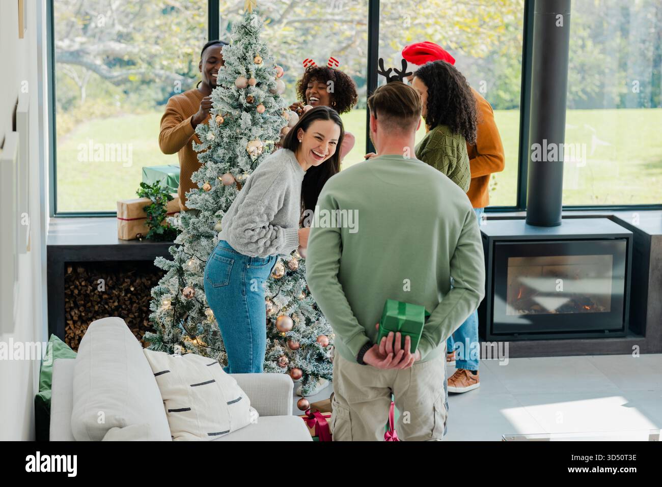 Amici diversi che decorano l'albero di Natale floccato nel soggiorno scambiando regali avvolti Foto Stock