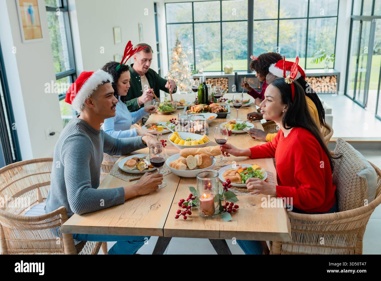 Diversi amici seduti intorno al tavolo da pranzo condividendo il pasto con bicchieri da vino e albero di Natale Foto Stock