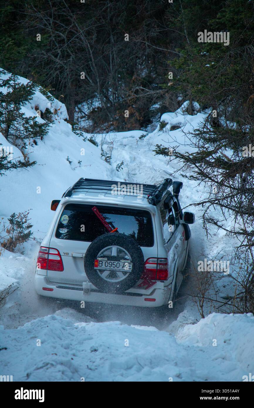 Naryn, Kirghizistan - 13 dicembre 2024: La vista posteriore di una Toyota Land Cruiser 100 bianca che guida su una stretta pista innevata, mescolando neve fresca in un'altra Foto Stock