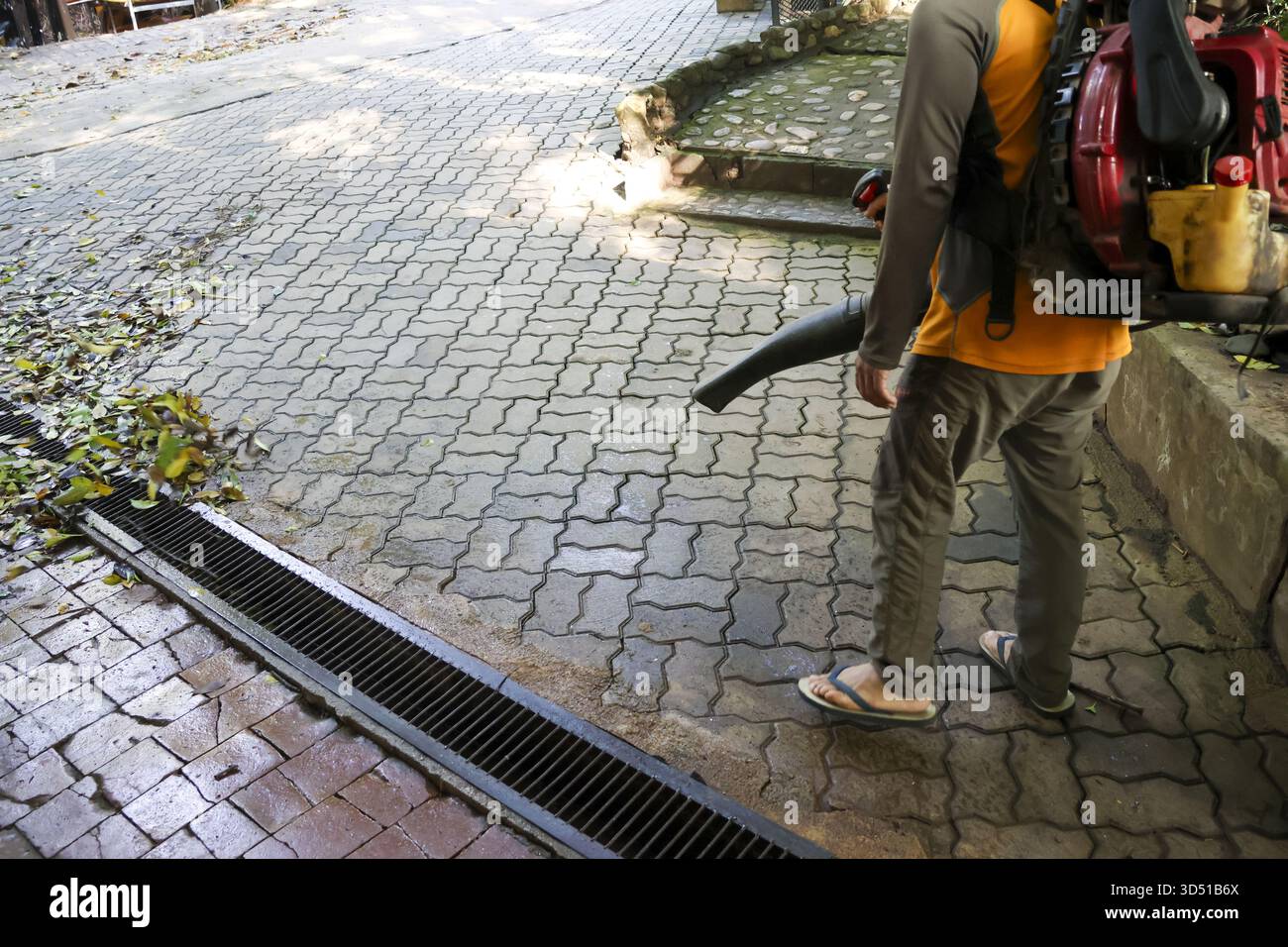 Operaio diligente che utilizza un soffiatore di foglie per pulire le foglie secche dal marciapiede in mattoni. Questo lavoro di manutenzione autunnale mostra una particolare attenzione al lavoro in cantiere Foto Stock