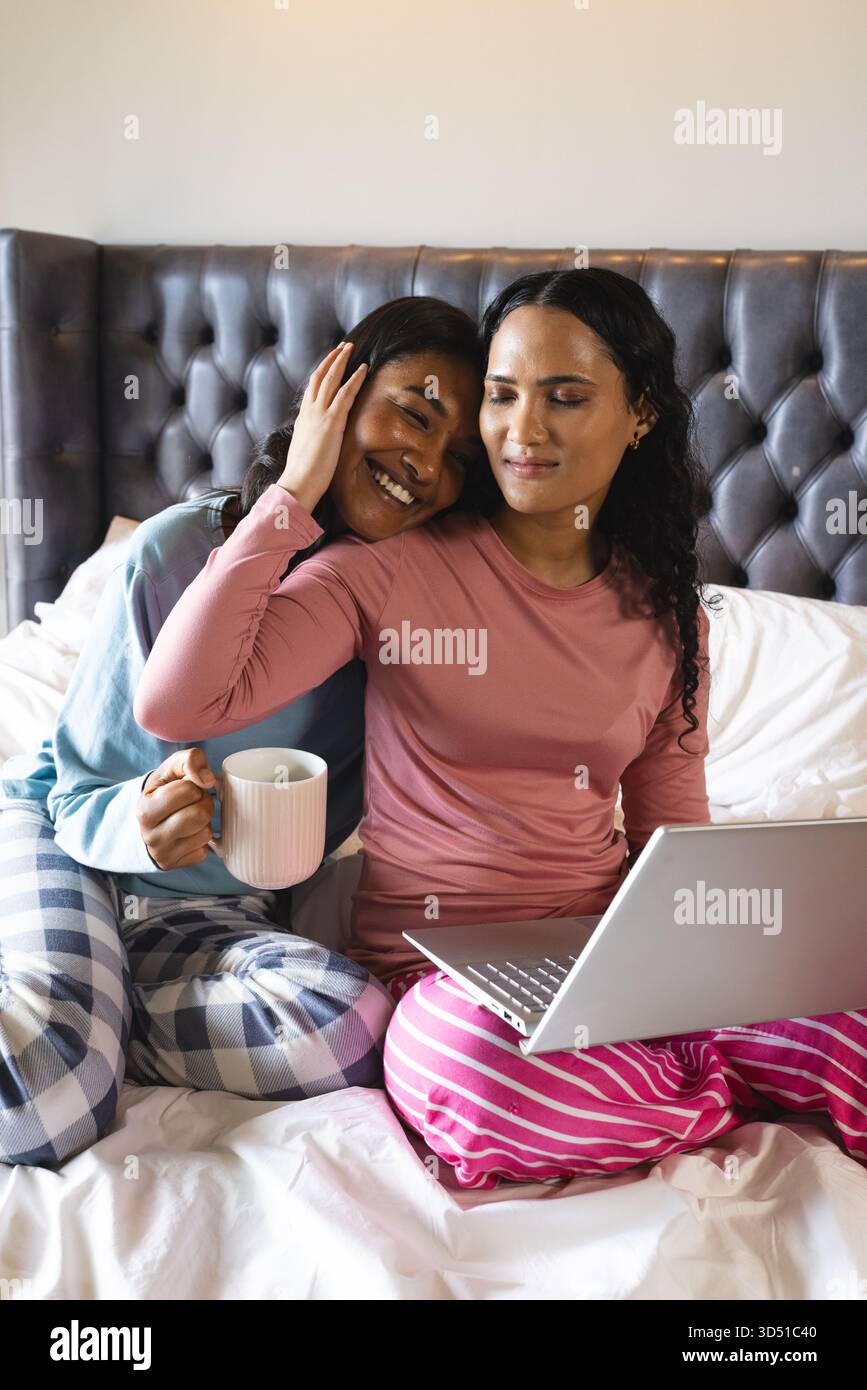 Coccolare diverse partner femminili che navigano su un laptop argentato sul letto in camera da letto, con tazza in ceramica bianca Foto Stock