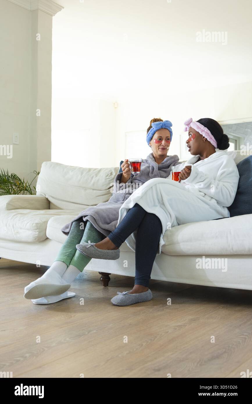 Rilassanti amiche diverse sorseggiando un tè alle erbe rosso sul divano nel soggiorno, con maschere facciali Foto Stock