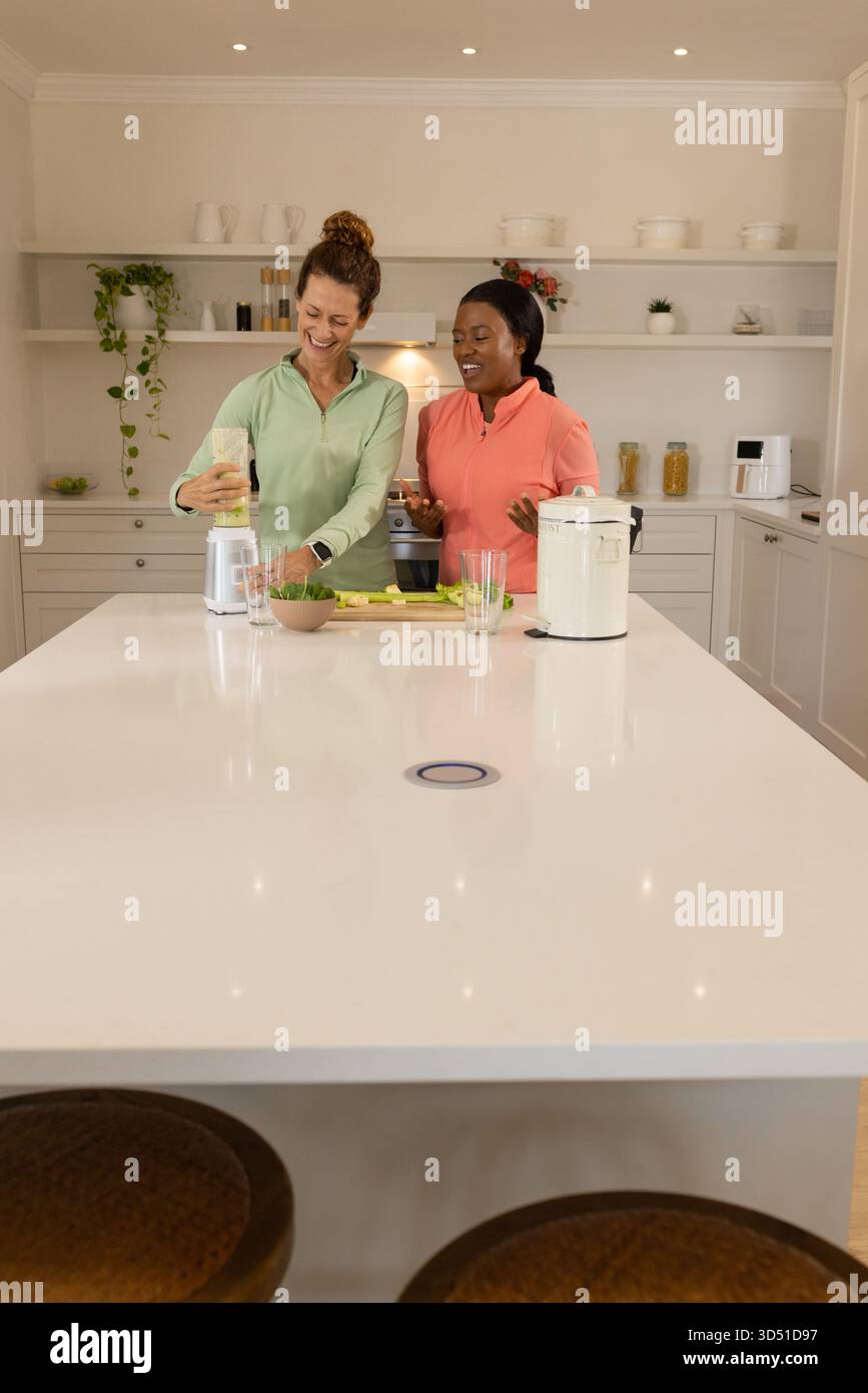 Sorridendo diverse amiche che mescolano frullati verdi nella cucina moderna, con frullatore e verdure Foto Stock