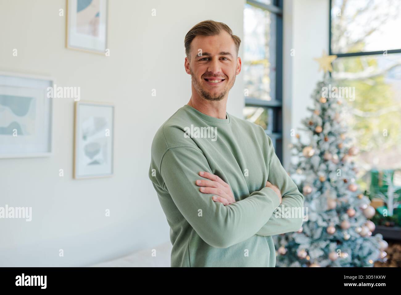 Uomo di trent'anni in piedi con le braccia incrociate, sorridente alla macchina fotografica in salotto con l'albero di Natale Foto Stock