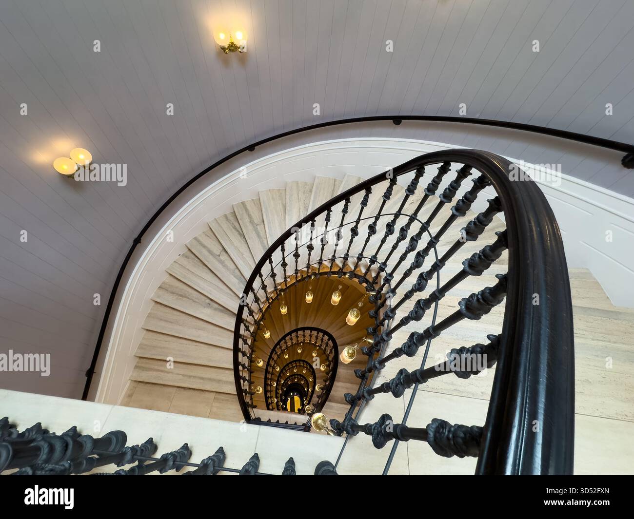 La scala a chiocciola mostra balaustre nere ornate gradini in marmo ringhiere curve e lampade calde che creano una prospettiva spettacolare verso il basso e un'elegante atmosfera storica Foto Stock