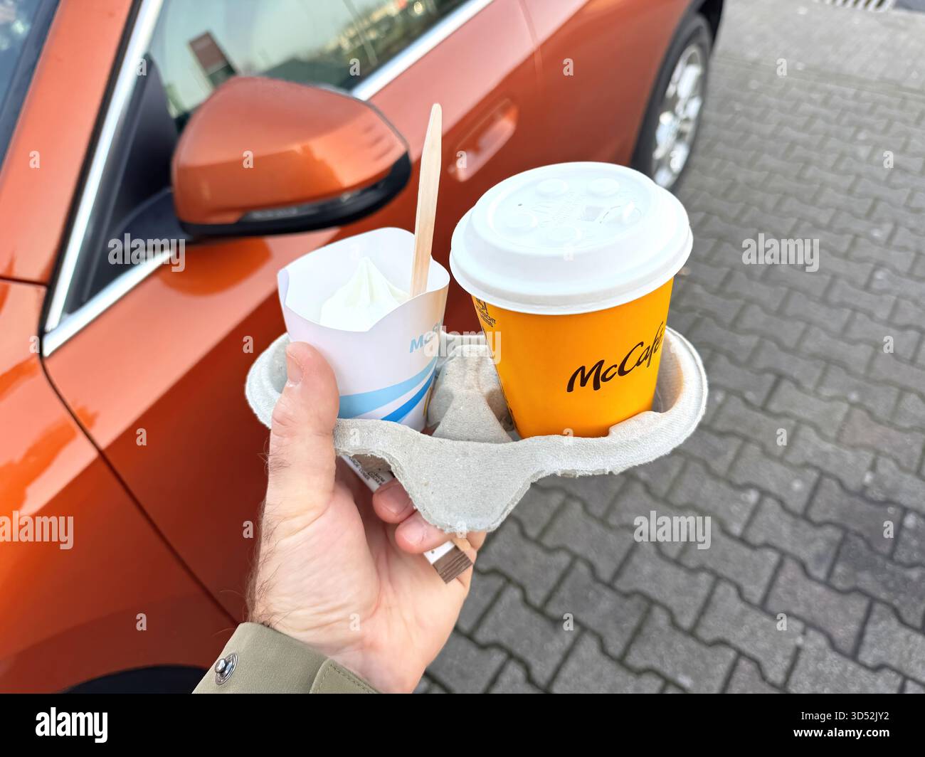 Francoforte, Germania - 1 aprile 2025: Uomo che tiene in mano una tazza di caffè McCafe e un gelato McFlurry in un vassoio di cartone vicino a un parcheggio elettrico BMW arancione Tahoe Foto Stock