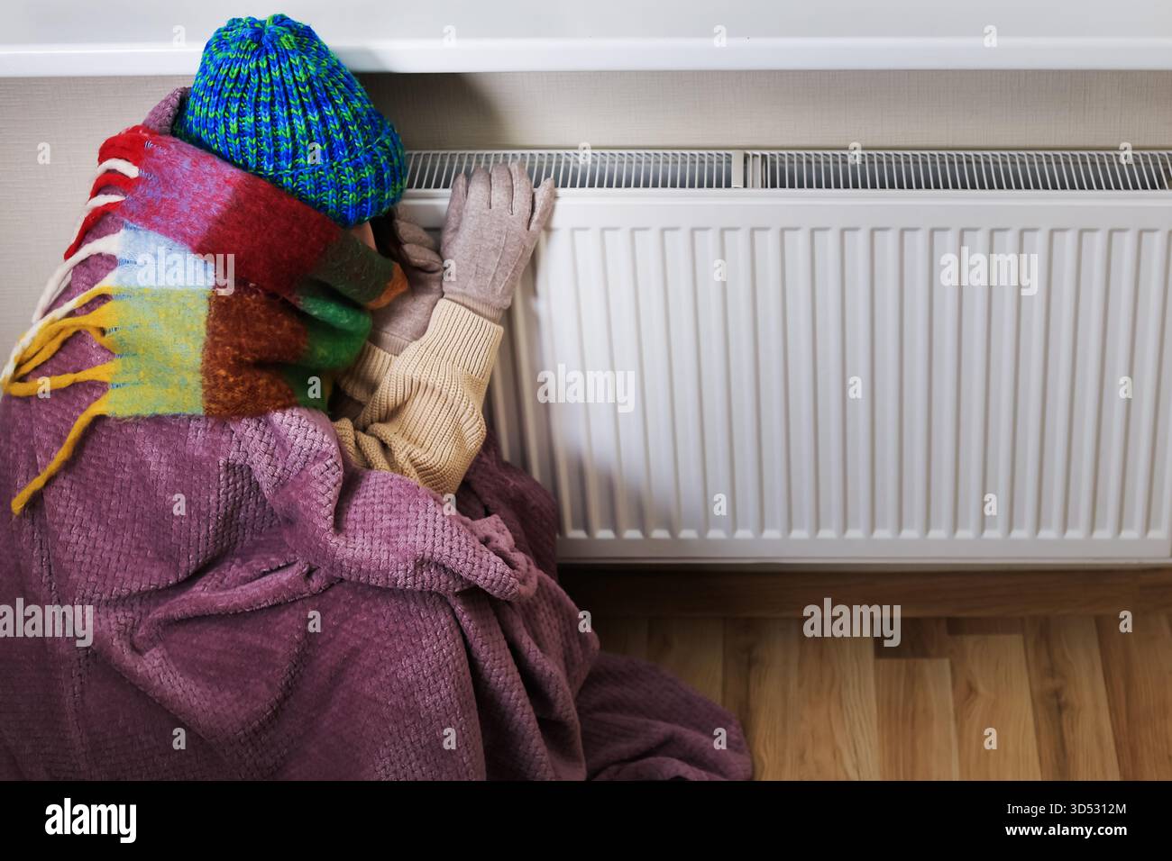 Una persona accarezzata da un cappello colorato, sciarpa e coperta, appoggiata su un radiatore bianco per tenersi al caldo durante il freddo o un problema di riscaldamento. Alta qualità Foto Stock