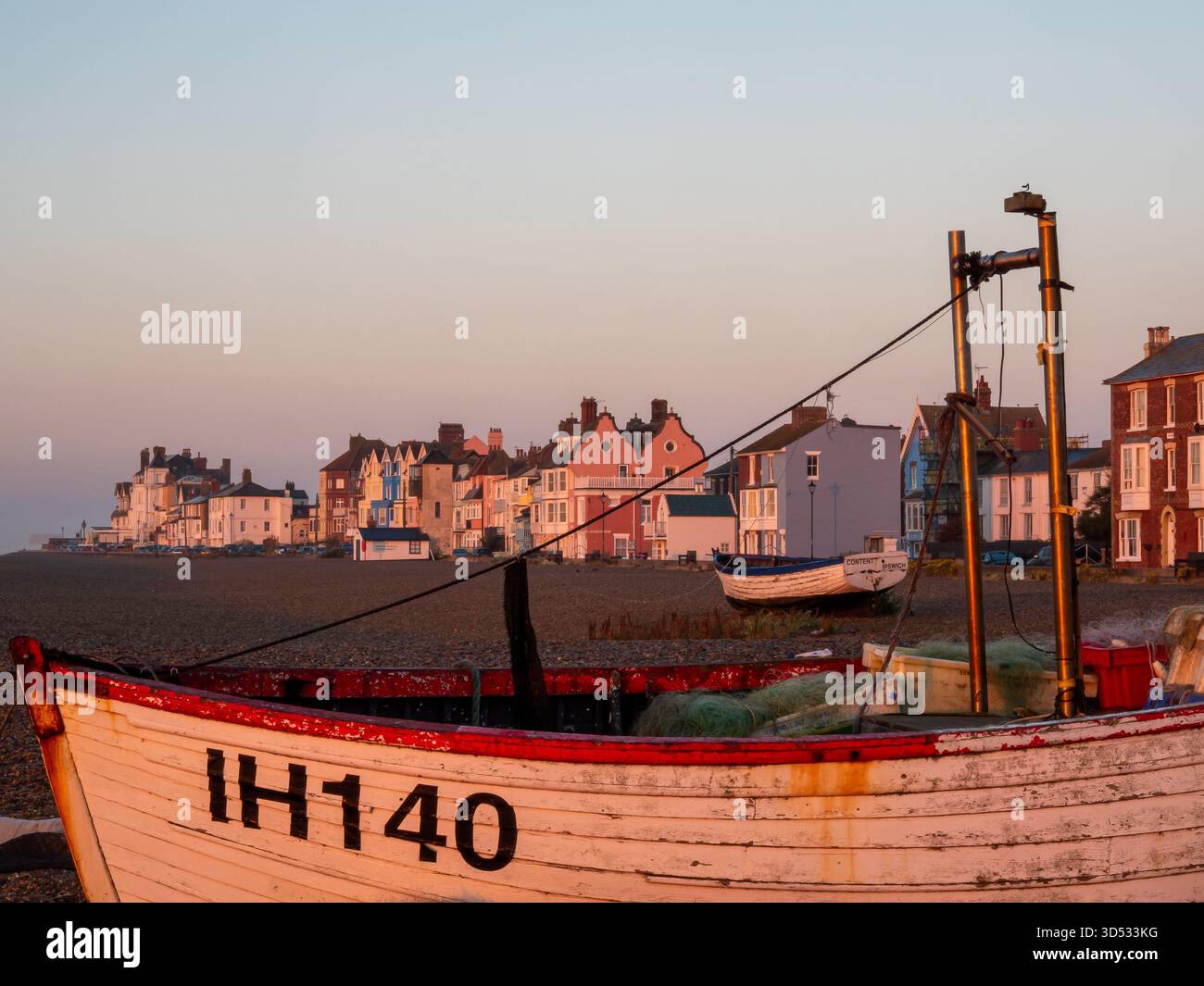 Tradizionale peschereccio di Aldeburgh che riposa sulla spiaggia di ciottoli con case color pastello sul lungomare illuminate da caldi colori dell'alba Foto Stock