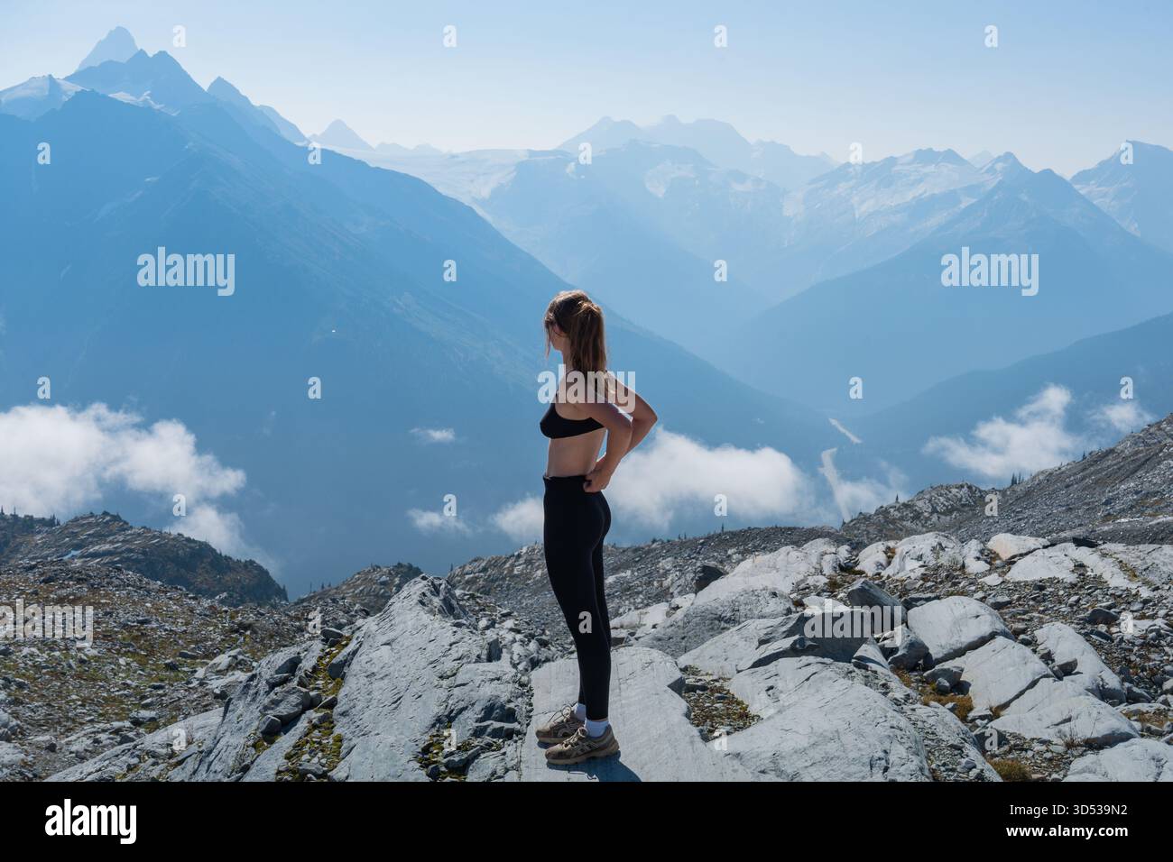 Giovane donna in abbigliamento sportivo che ammira la vista sulle montagne da una cima rocciosa. Glacier National Park, British Columbia Foto Stock