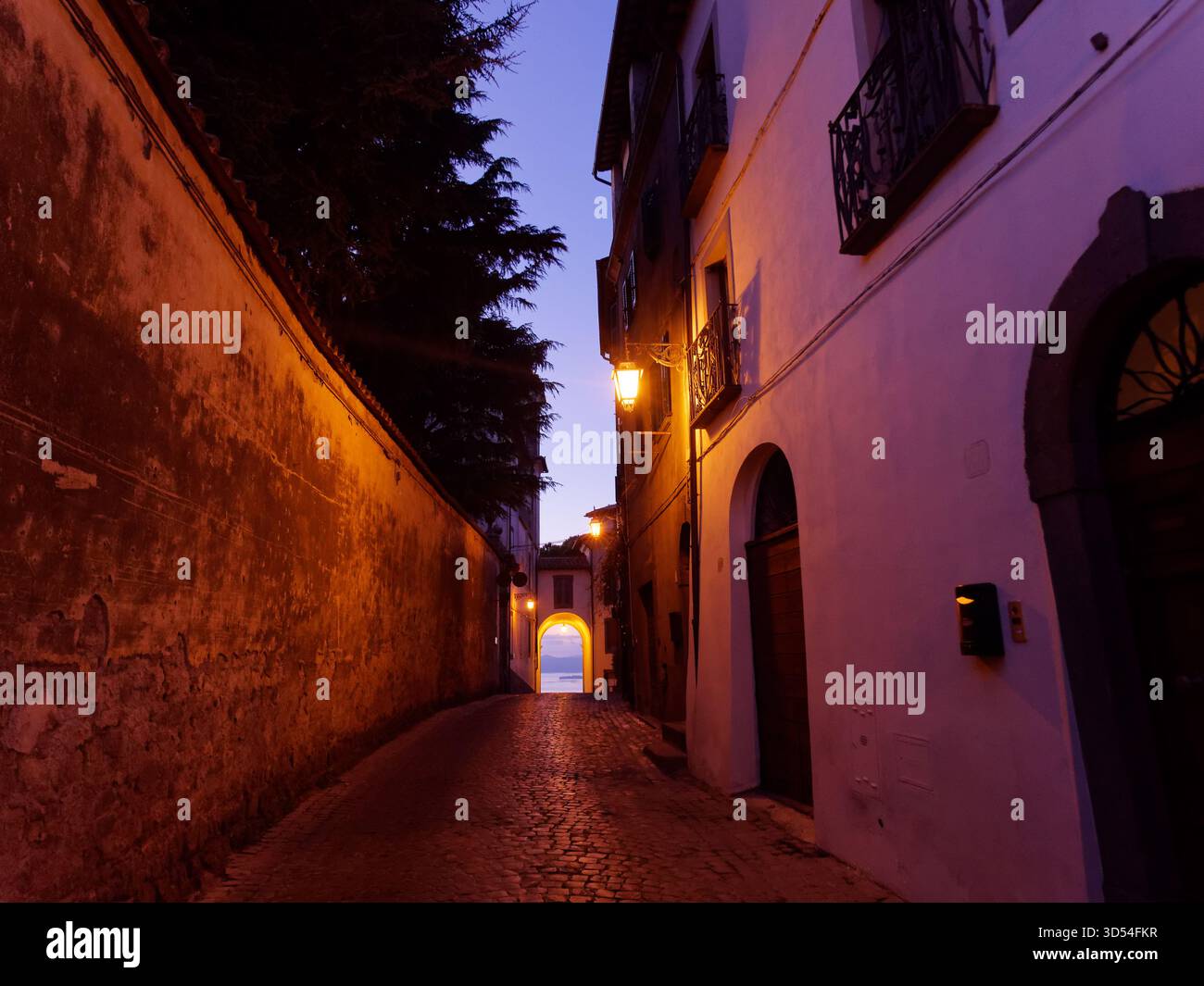 Stretta strada acciottolata con alte mura e arcata verso il lago di Bolsena al crepuscolo mentre si illumina un semaforo. Montefiascone, Lazio, Italia. 12 novembre 2025 Foto Stock