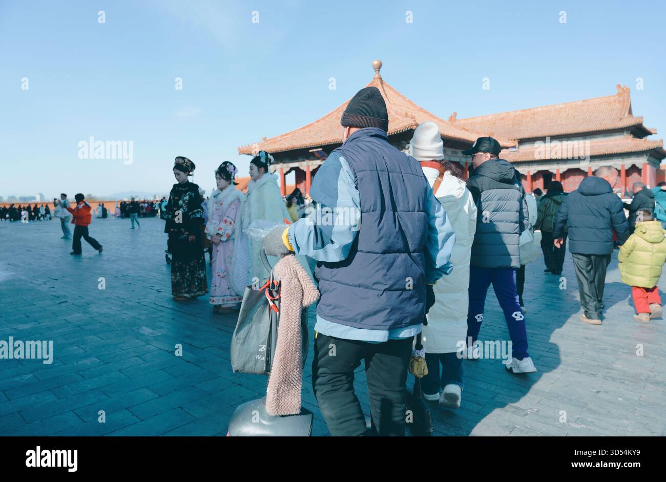 Le persone a strati invernali camminano attraverso una piazza della città Proibita mentre le donne in un hanfu tradizionale posano vicino ai tetti del palazzo sotto un luminoso cielo di Pechino Foto Stock