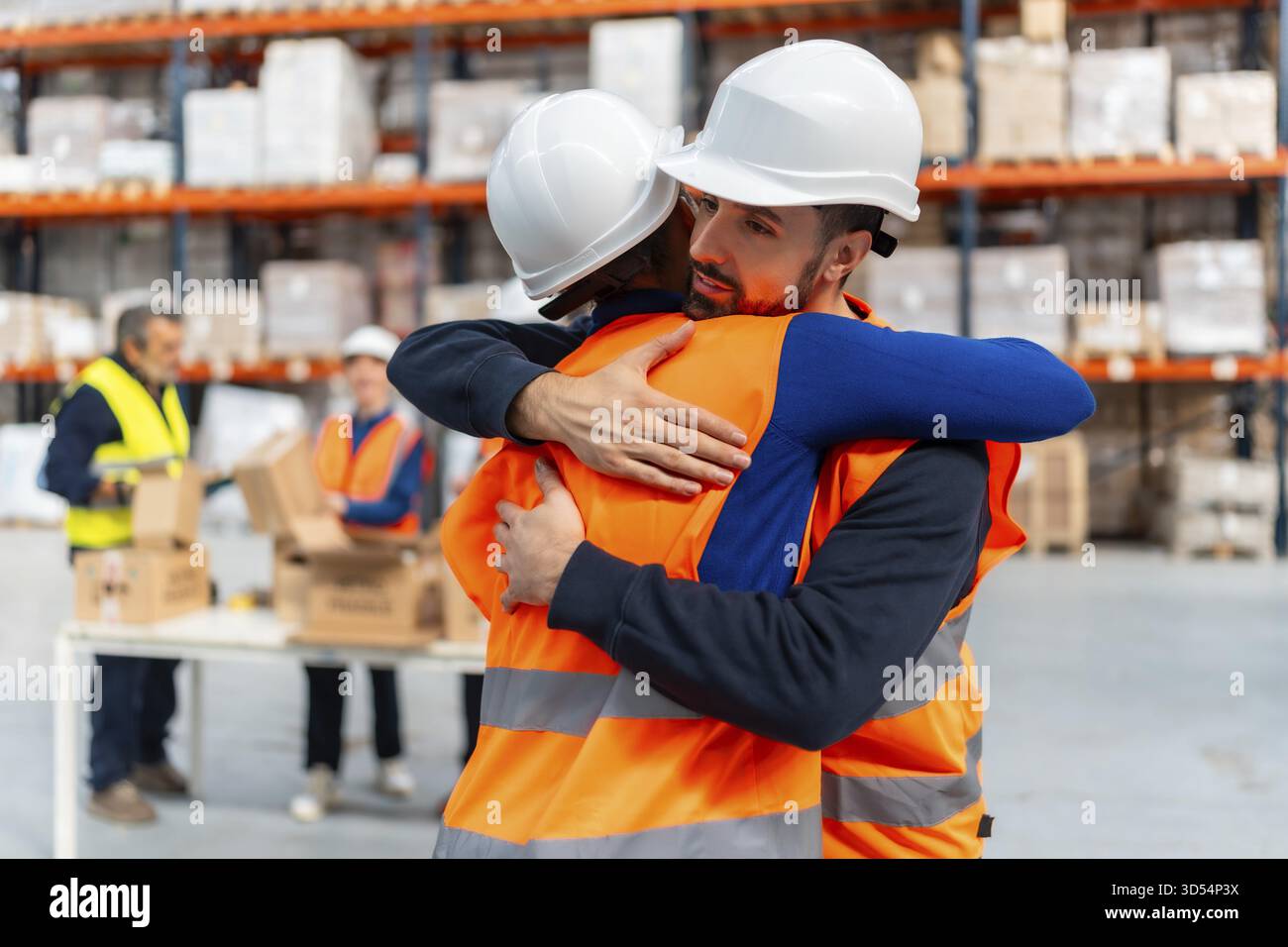 Due diversi operatori di magazzino che indossano giubbotti di sicurezza e cappelli rigidi che abbracciano in una struttura logistica, simboleggiano l'unità, il supporto e un forte senso di c Foto Stock