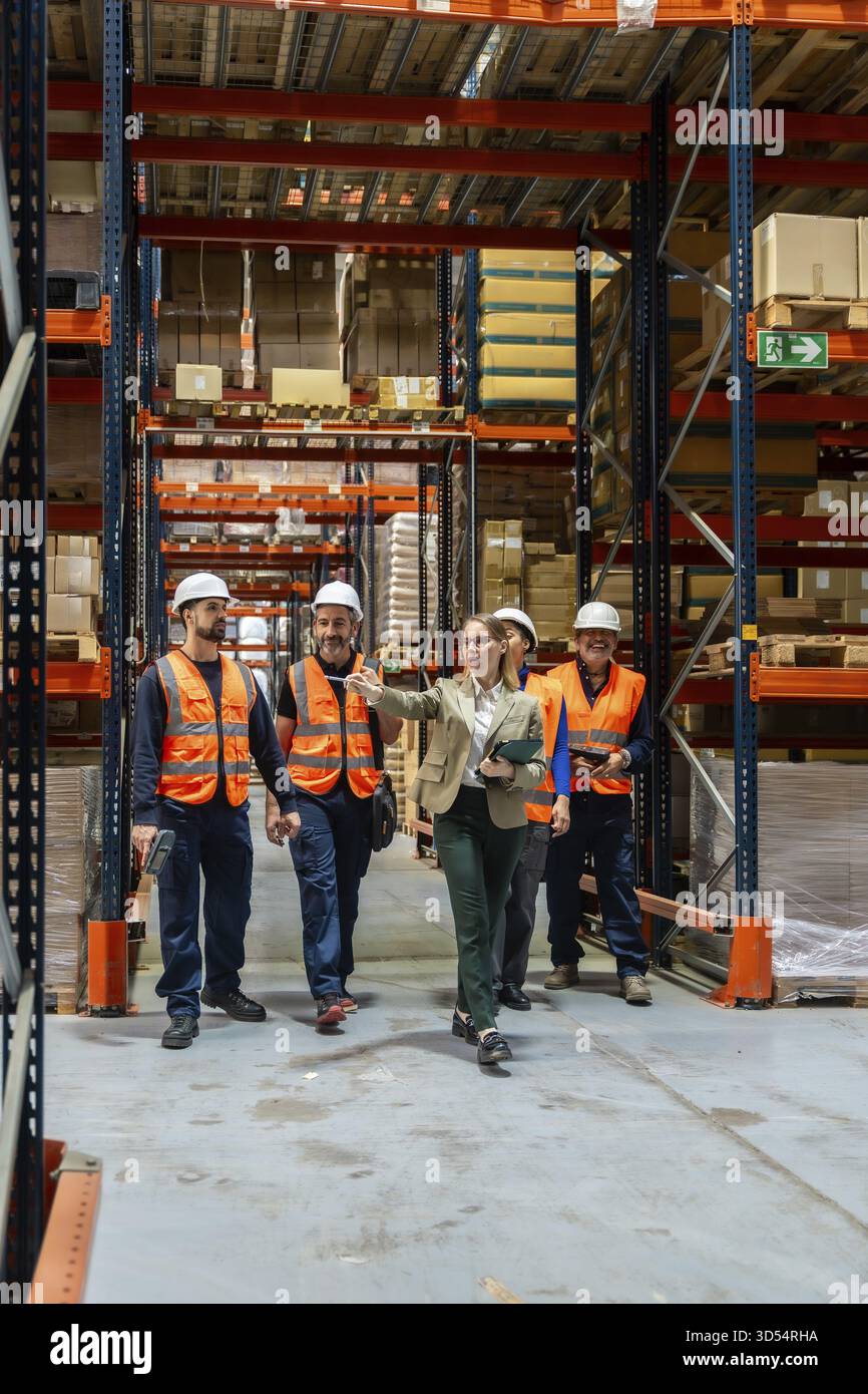 Team diversificato di professionisti della logistica e di addetti al magazzino che discutono della gestione dell'inventario e delle procedure operative sicure mentre attraversano un Foto Stock