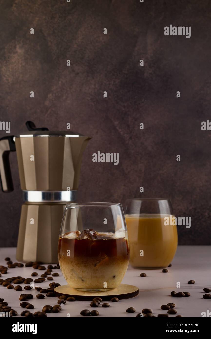Accanto a due bicchieri è presente una caffettiera riempita di caffè freddo e latte. Intorno a loro si trovano chicchi di caffè sparsi su una superficie scura, creando un Foto Stock
