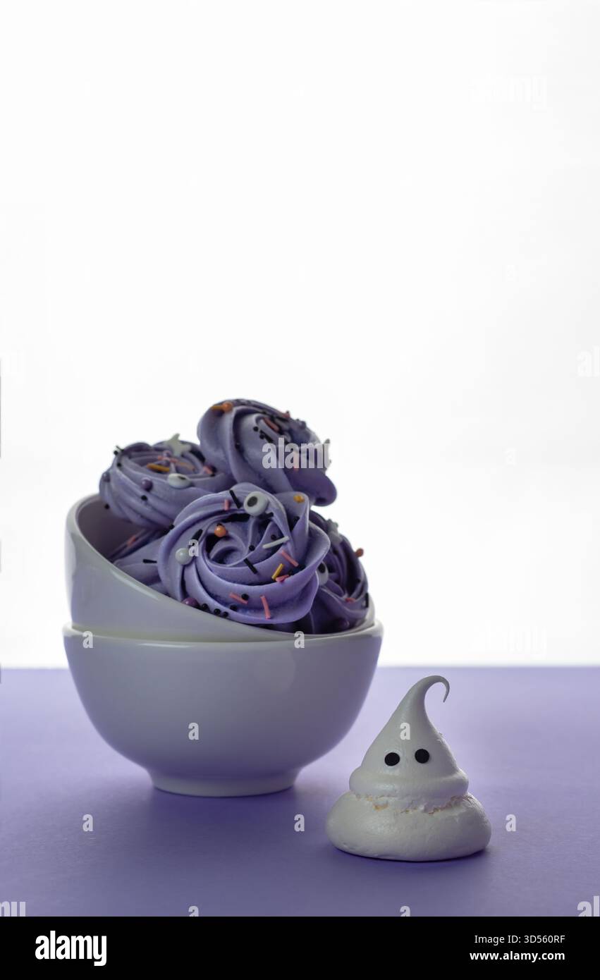 Una ciotola contiene diversi turni di meringa viola dalla forma splendida e ricoperti di sprinkles, mentre una piccola meringa a forma di fantasma si trova nelle vicinanze su un surf leggero Foto Stock