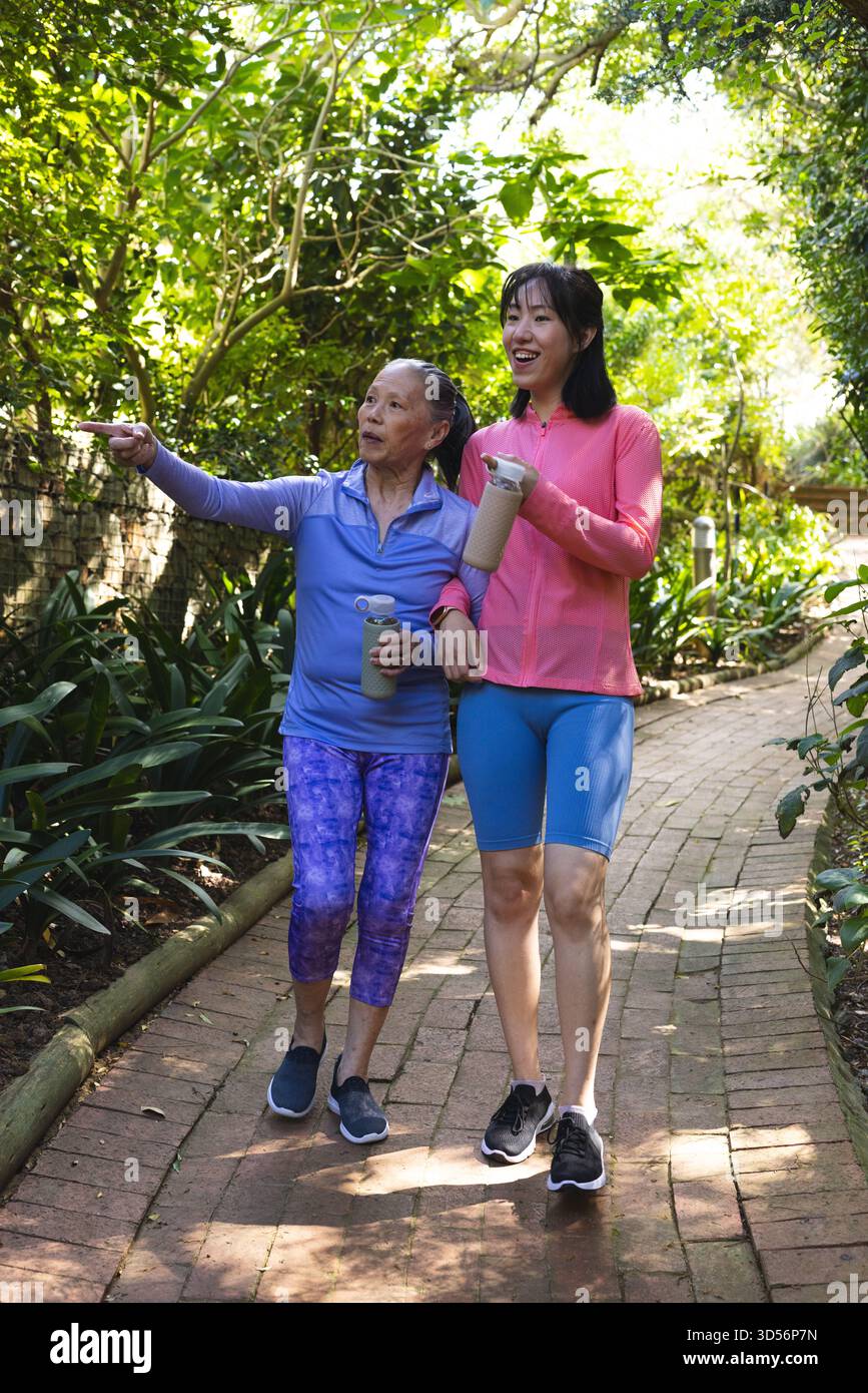 Madre e figlia asiatiche che camminano con un braccio sul sentiero in mattoni, tenendo in mano una bottiglia d'acqua e un bicchiere Foto Stock