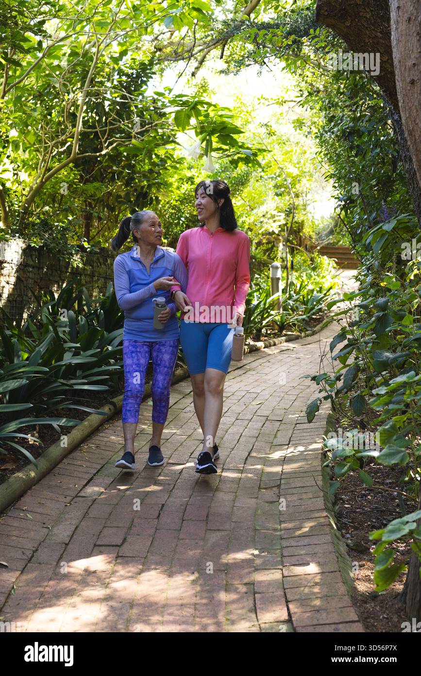 Camminando tra la madre e la figlia asiatica cinese con un braccio sul sentiero in mattoni in giardino, tenendo bottiglie d'acqua Foto Stock