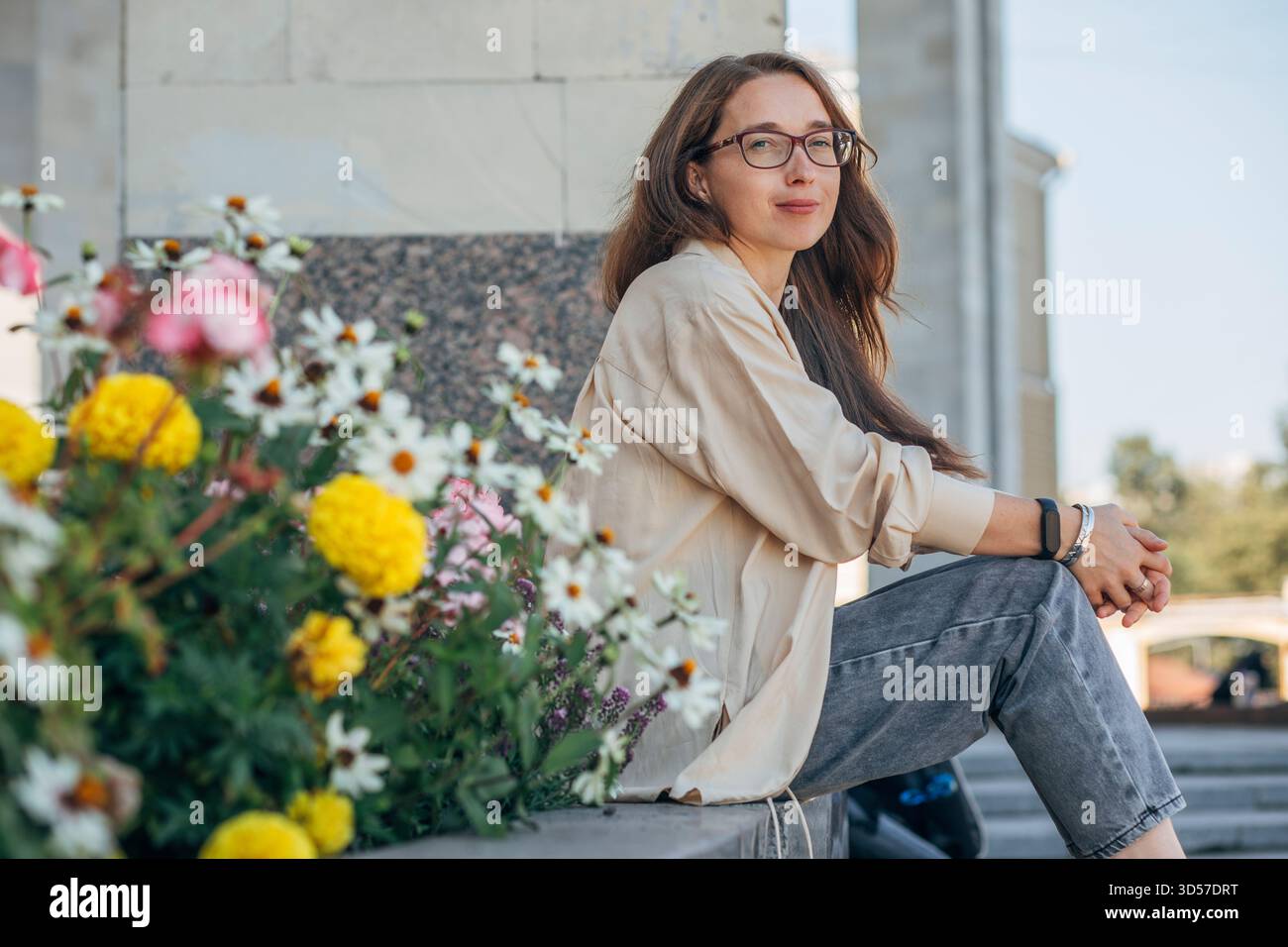 Donna con occhiali seduti accanto ai fiori della città, ritratto rilassato all'aperto Foto Stock