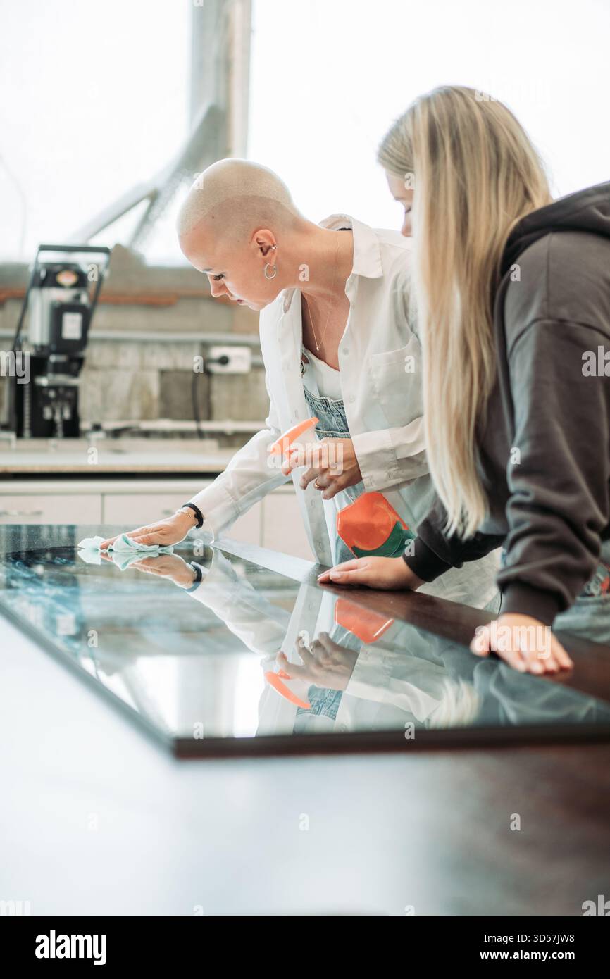 Donna calva che pulisce la superficie in vetro con flacone spray in officina Foto Stock