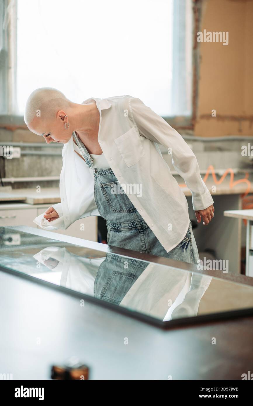 Donna con testa rasata in tuta che ispeziona i pannelli di vetro in officina Foto Stock
