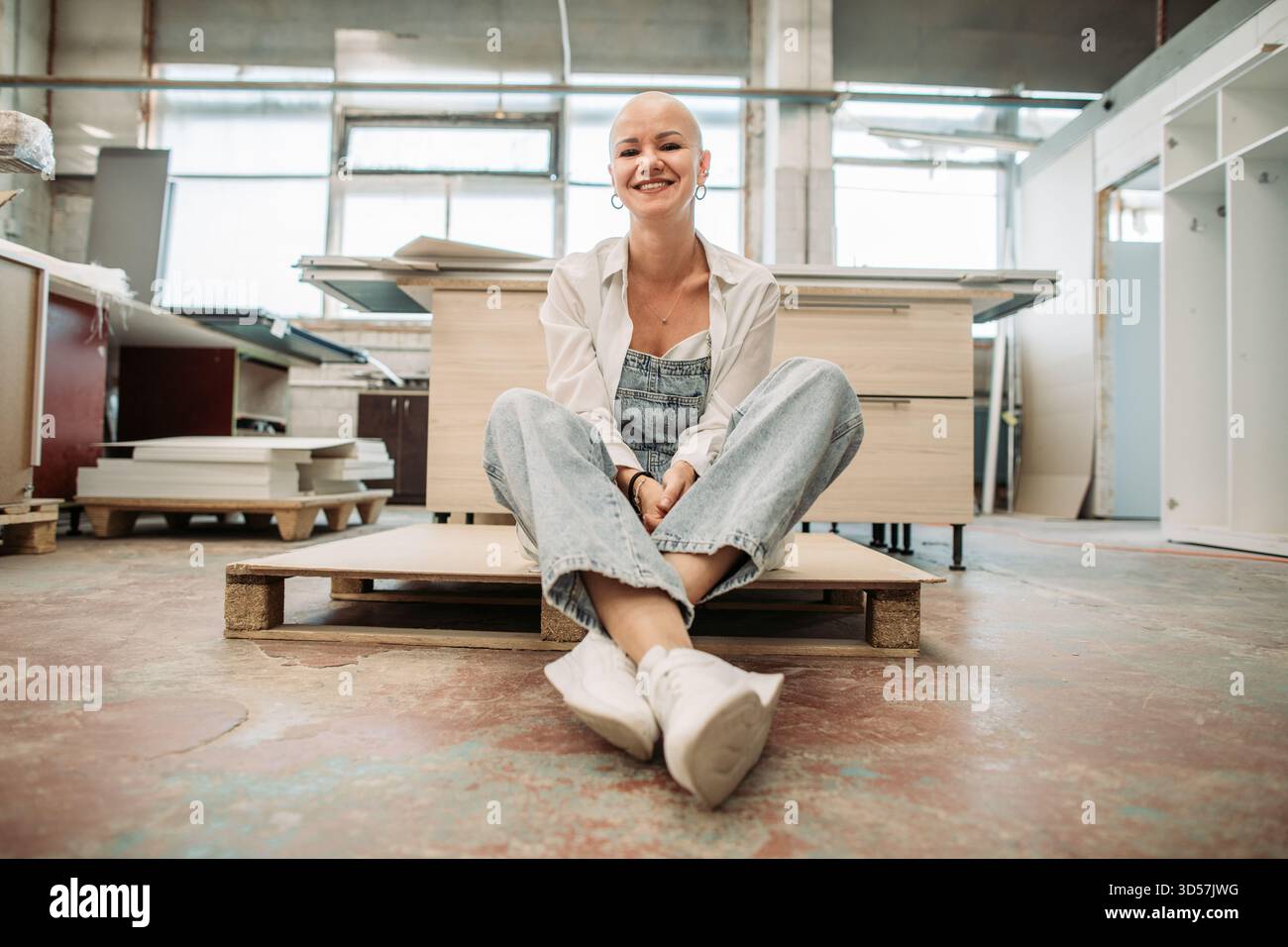 Donna calva sorridente in tuta seduta su pallet presso un'officina di mobili Foto Stock