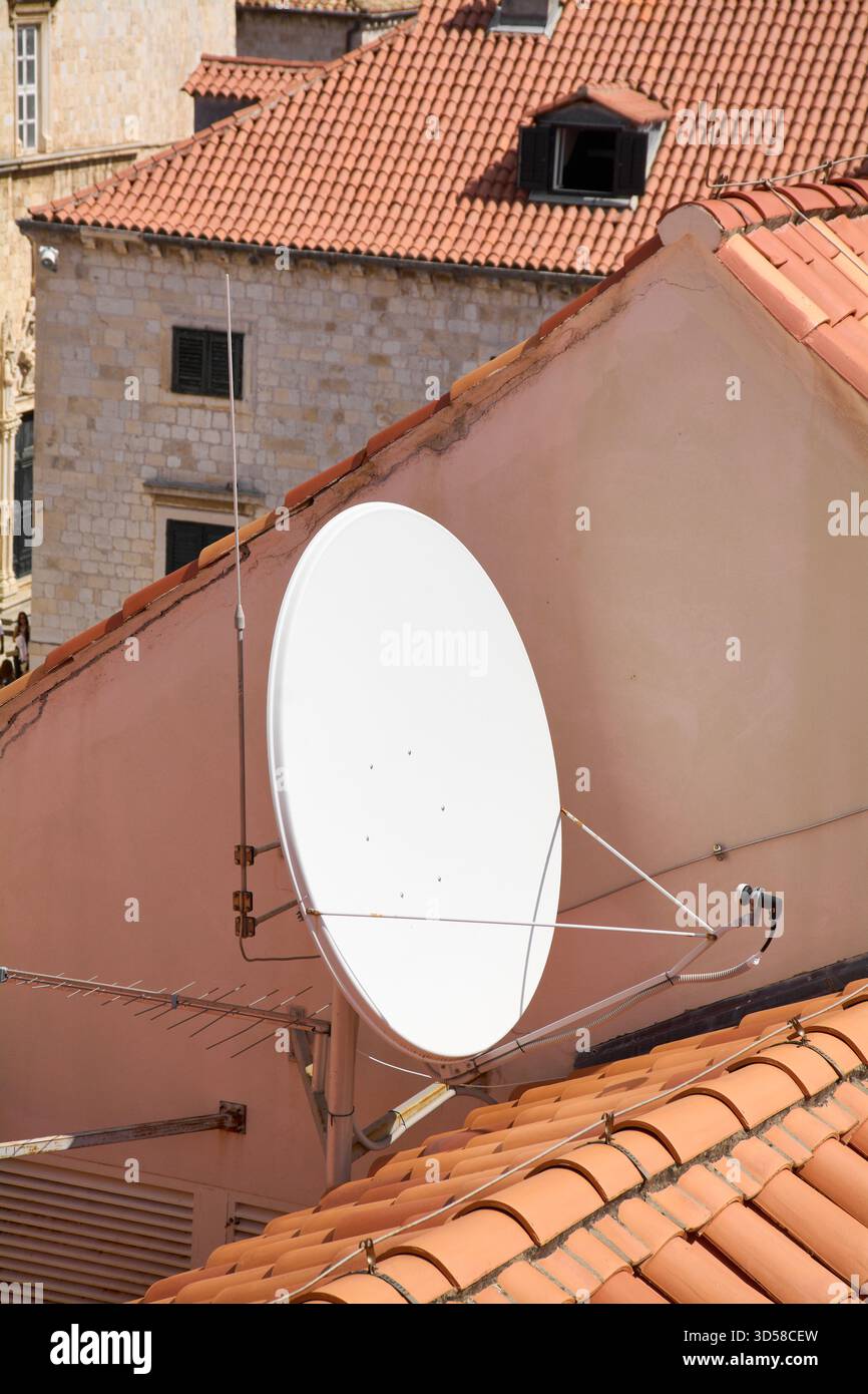 Particolare di una parabola parabolica satellitare per la comunicazione e la televisione montata sulla parete di un vecchio edificio con tetti di tegole rosse. Il vecchio incontra il nuovo idiota Foto Stock