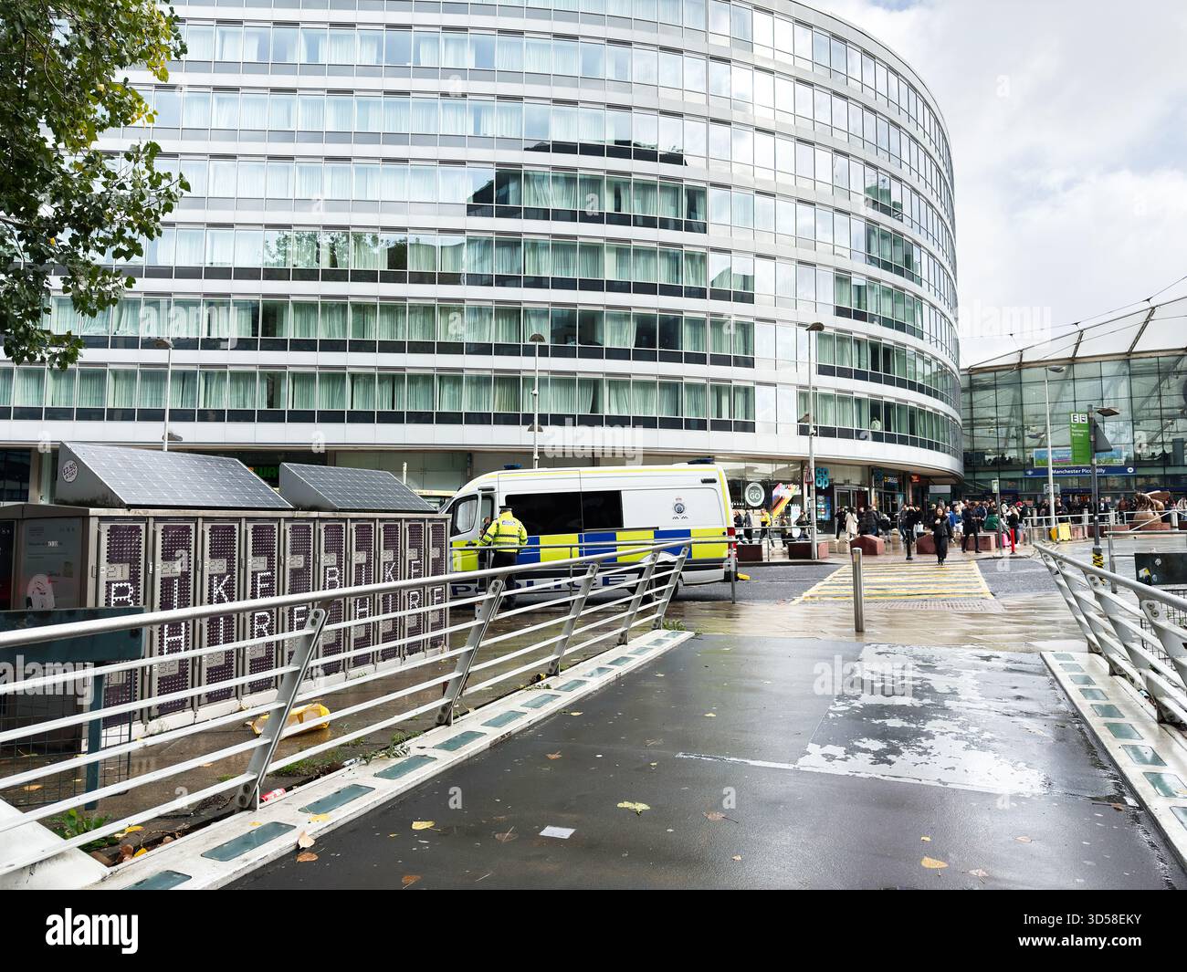 Manchester, Regno Unito - 4 ottobre 2025: Vivace scena urbana fuori da un edificio curvo in vetro della stazione di Manchester Piccadilly con ambulanza e pedoni. Foto Stock