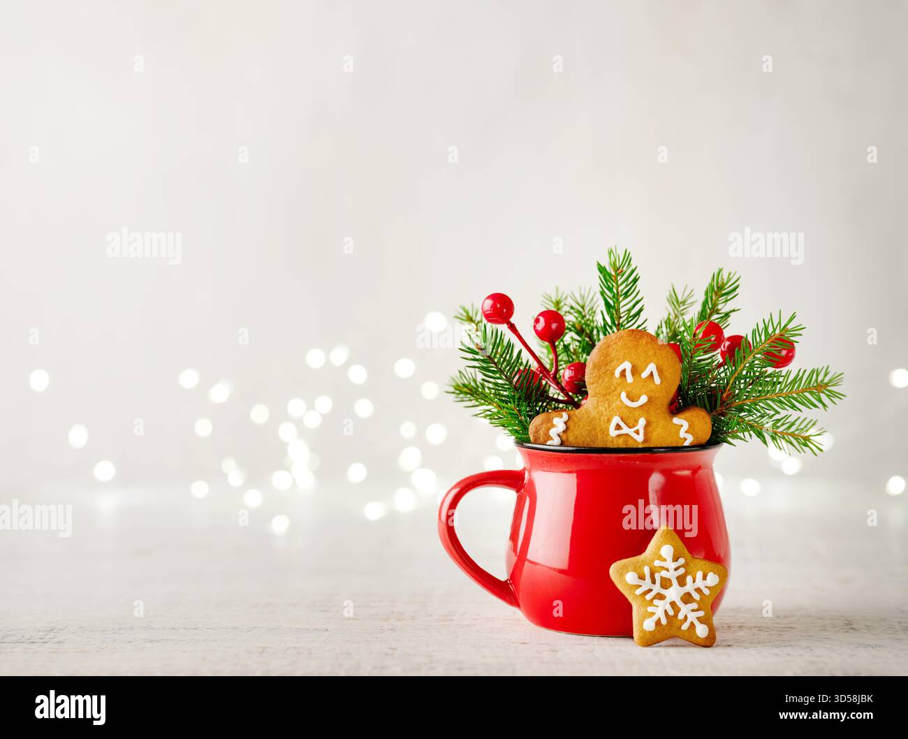Uomo di pan di zenzero di Natale in tazza rossa con biscotti caldi alla cioccolata con decorazioni, rami dell'albero di Natale, regali e giocattoli su sfondo in legno con bokeh garland. Elemento di design del biglietto di Natale. Foto Stock