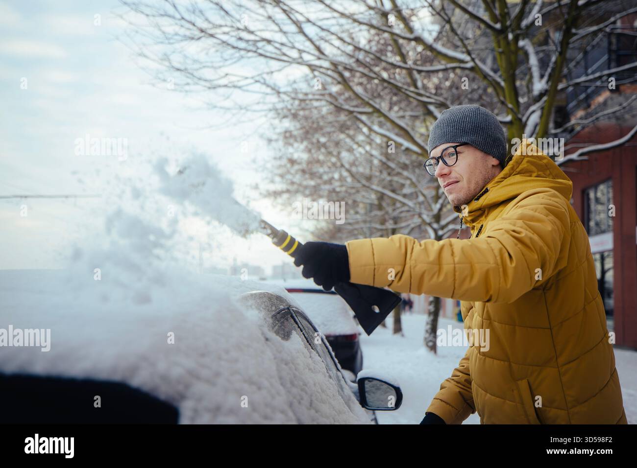 Uomo con abbigliamento invernale che pulisce la neve dall'auto parcheggiata in una strada residenziale della città durante il freddo mattino. Scena che mostra la routine invernale di tutti i giorni, meteo co Foto Stock