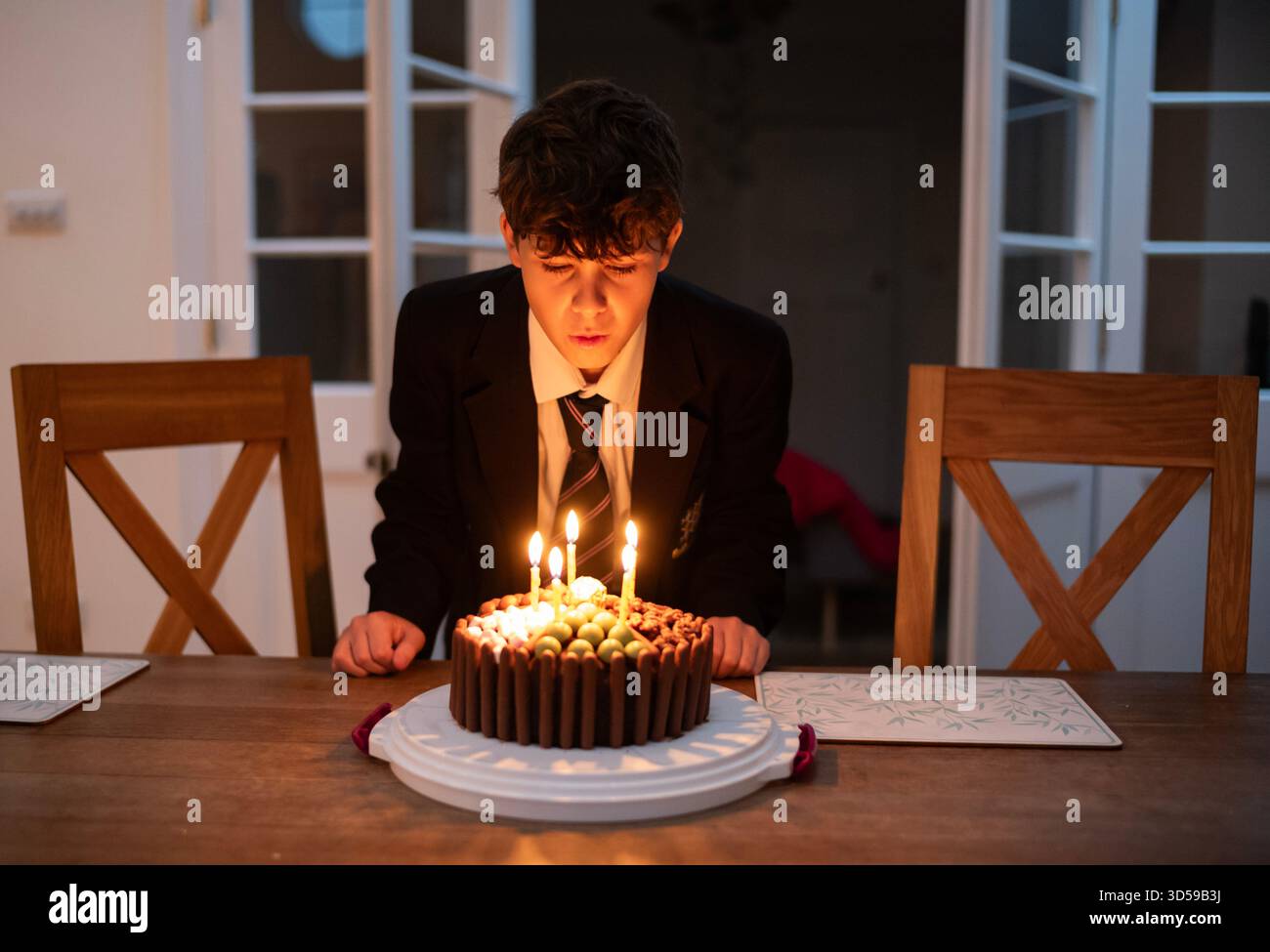 Un ragazzo di 12 anni fa fa saltare le candele sulla torta di compleanno indossando l'uniforme scolastica, dopo essere tornato a casa da scuola, Inghilterra, Regno Unito Foto Stock