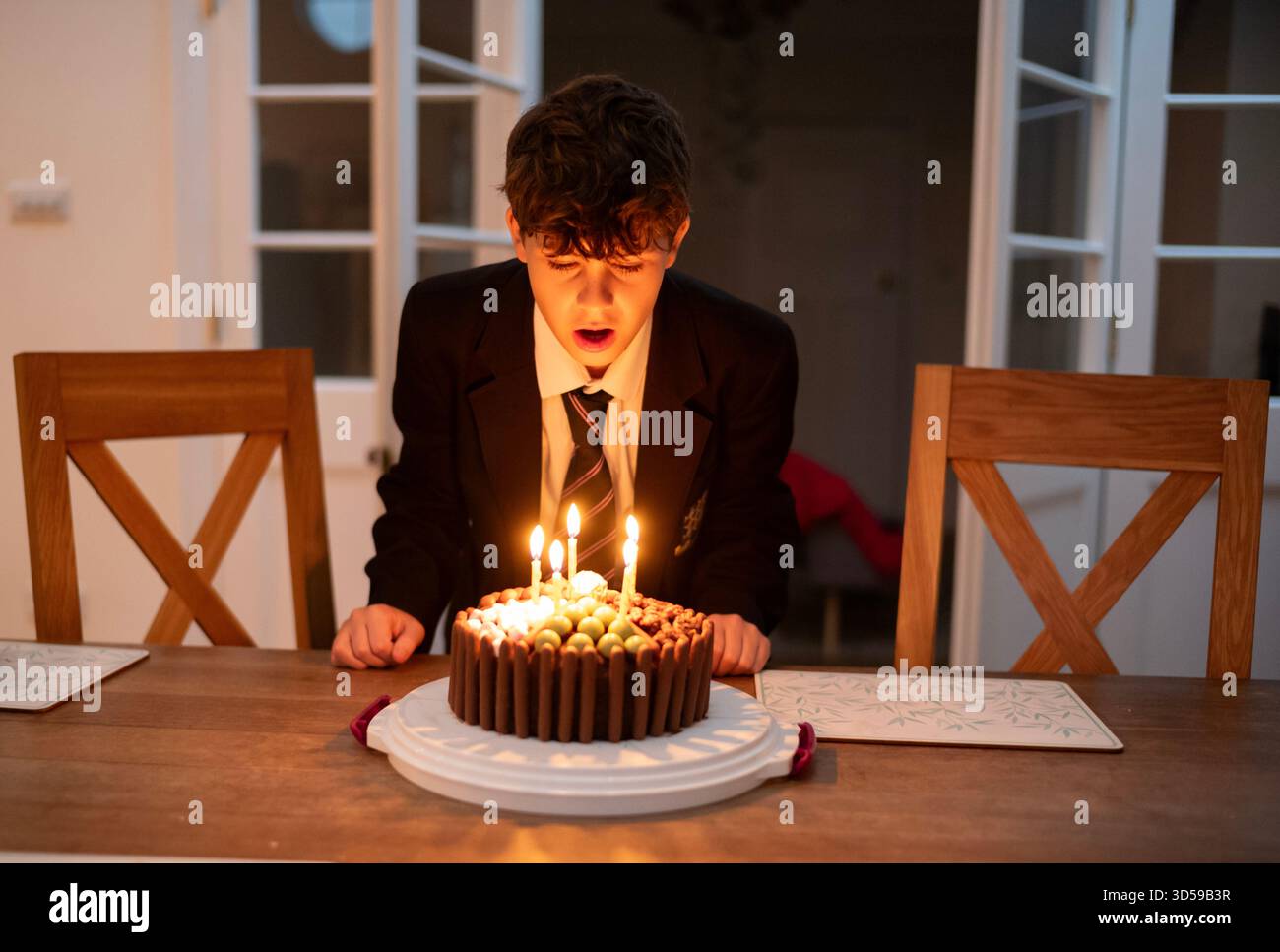 Un ragazzo di 12 anni fa fa saltare le candele sulla torta di compleanno indossando l'uniforme scolastica, dopo essere tornato a casa da scuola, Inghilterra, Regno Unito Foto Stock