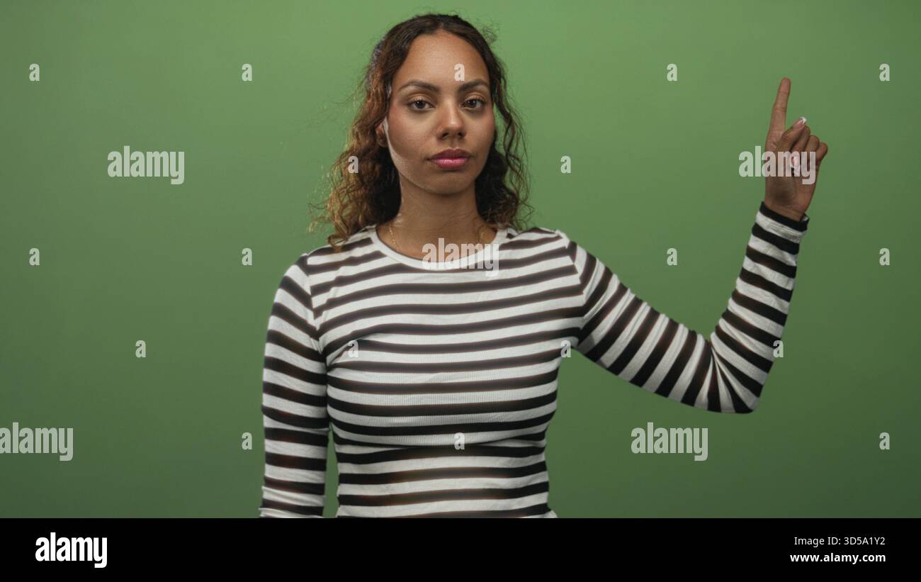 La donna punta il dito indice verso l'alto in studio indossando una camicia a righe sullo sfondo verde; massima tranquillità. Foto Stock