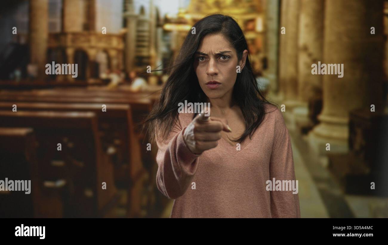 Donna che cammina con la testa inclinata vicino ai picchi di legno all'interno dell'edificio della chiesa, indossa un maglione rosa con scollo a V e un piccolo orecchino; dubbio religioso. Foto Stock