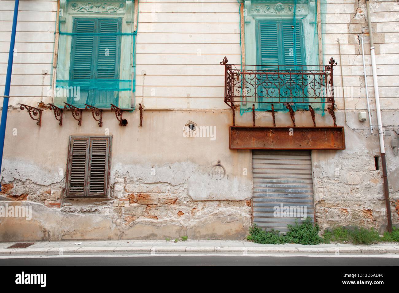 La facciata di un edificio intempestivo presenta persiane turchesi, un balcone decorativo in ferro, ringhiere arrugginite e un muro sbriciolato. L'esterno logoro evoca l'urba Foto Stock