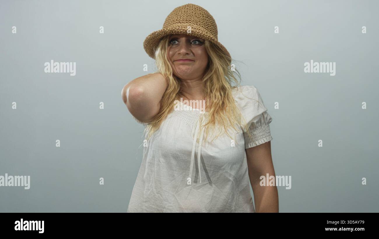 Donna con la mano sul collo, cappello di paglia e camicetta bianca in studio blu pallido, che guarda lateralmente con postura casual; contemplazione. Foto Stock