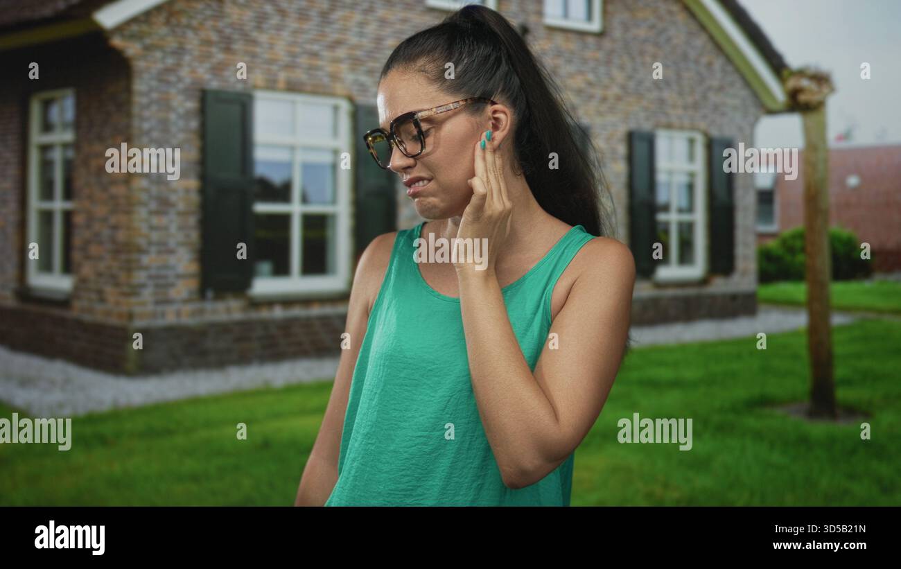 Donna con canotta verde e occhiali tiene la guancia con la mano per il mal di denti di fronte all'ingresso di un edificio in mattoni marrone; dolore. Foto Stock