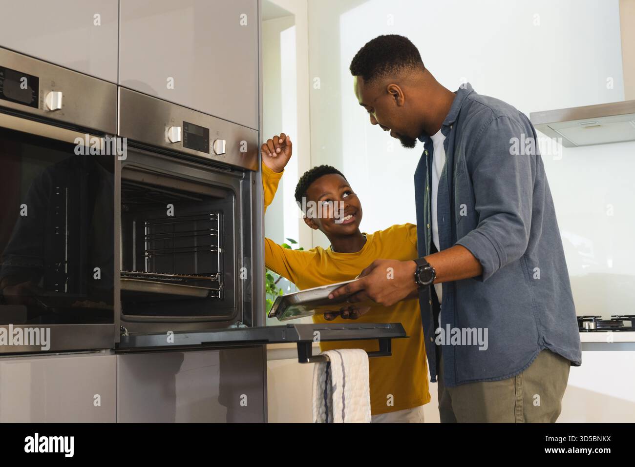Padre e figlio afroamericani che preparano la pizza in una cucina moderna con vassoio da forno e forno Foto Stock