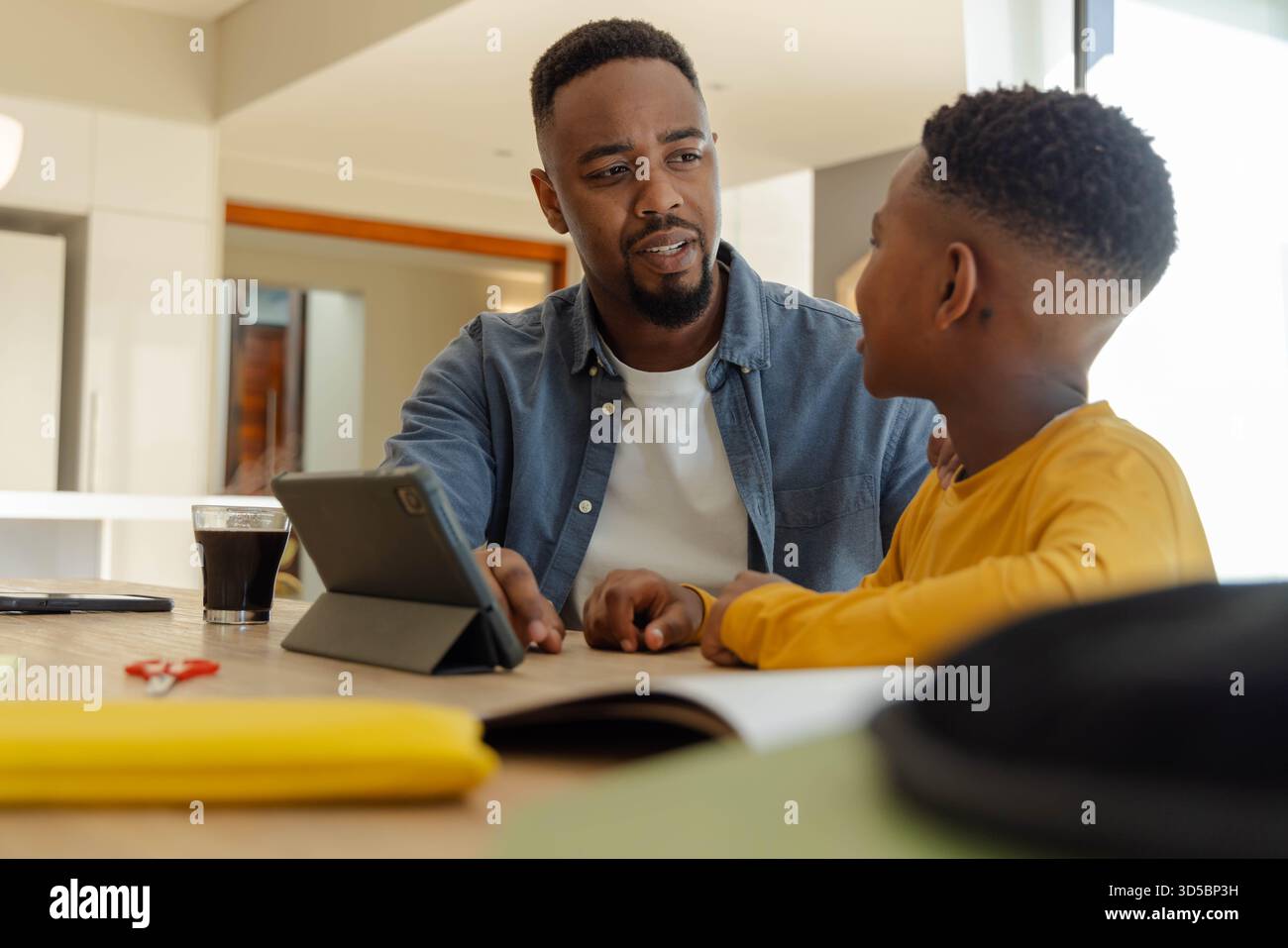 Padre e figlio discutono i compiti al tavolo della cucina con tablet e libri Foto Stock