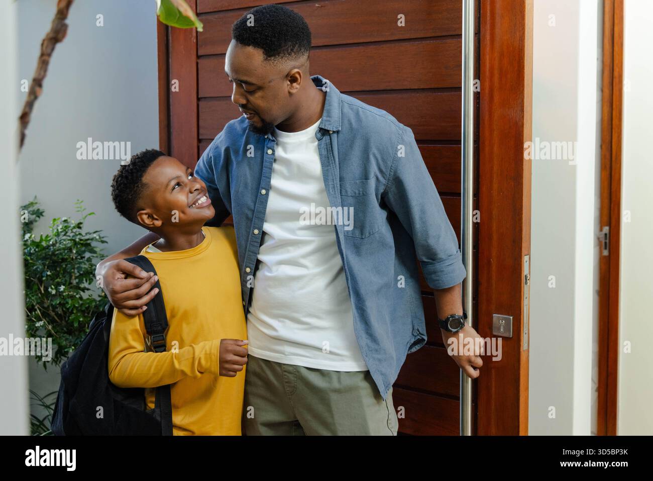 Padre e figlio sorridenti, abbracciati a casa, pronti per la scuola con lo zaino Foto Stock