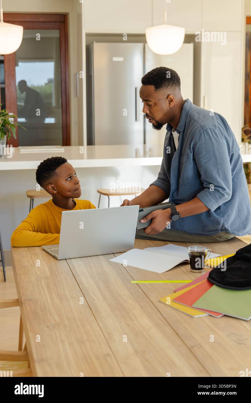Padre che aiuta il figlio con i compiti usando il laptop al tavolo della cucina, entrambi concentrati Foto Stock