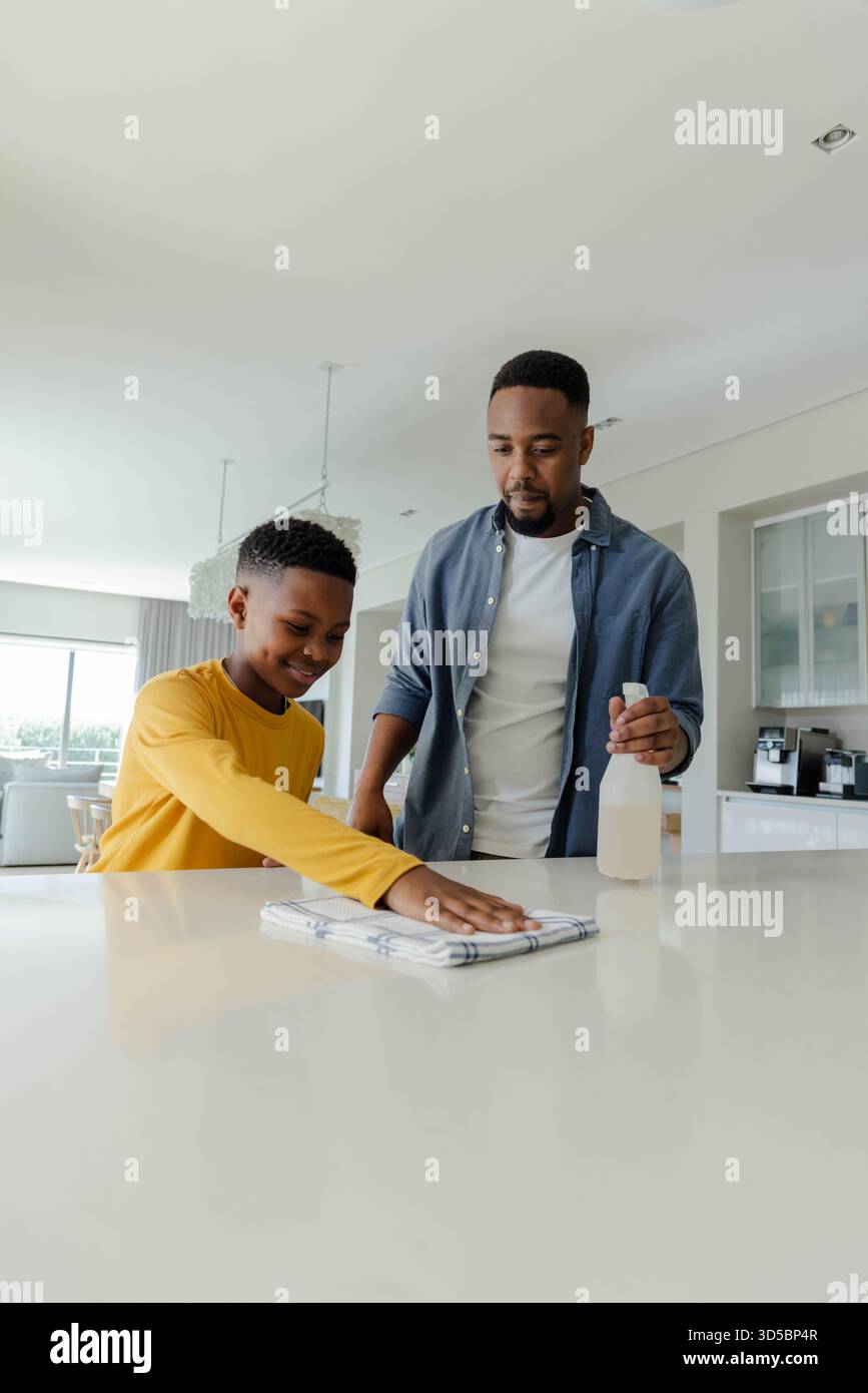 Padre e figlio afroamericani puliscono insieme il bancone della cucina, legano e sorridono a casa Foto Stock