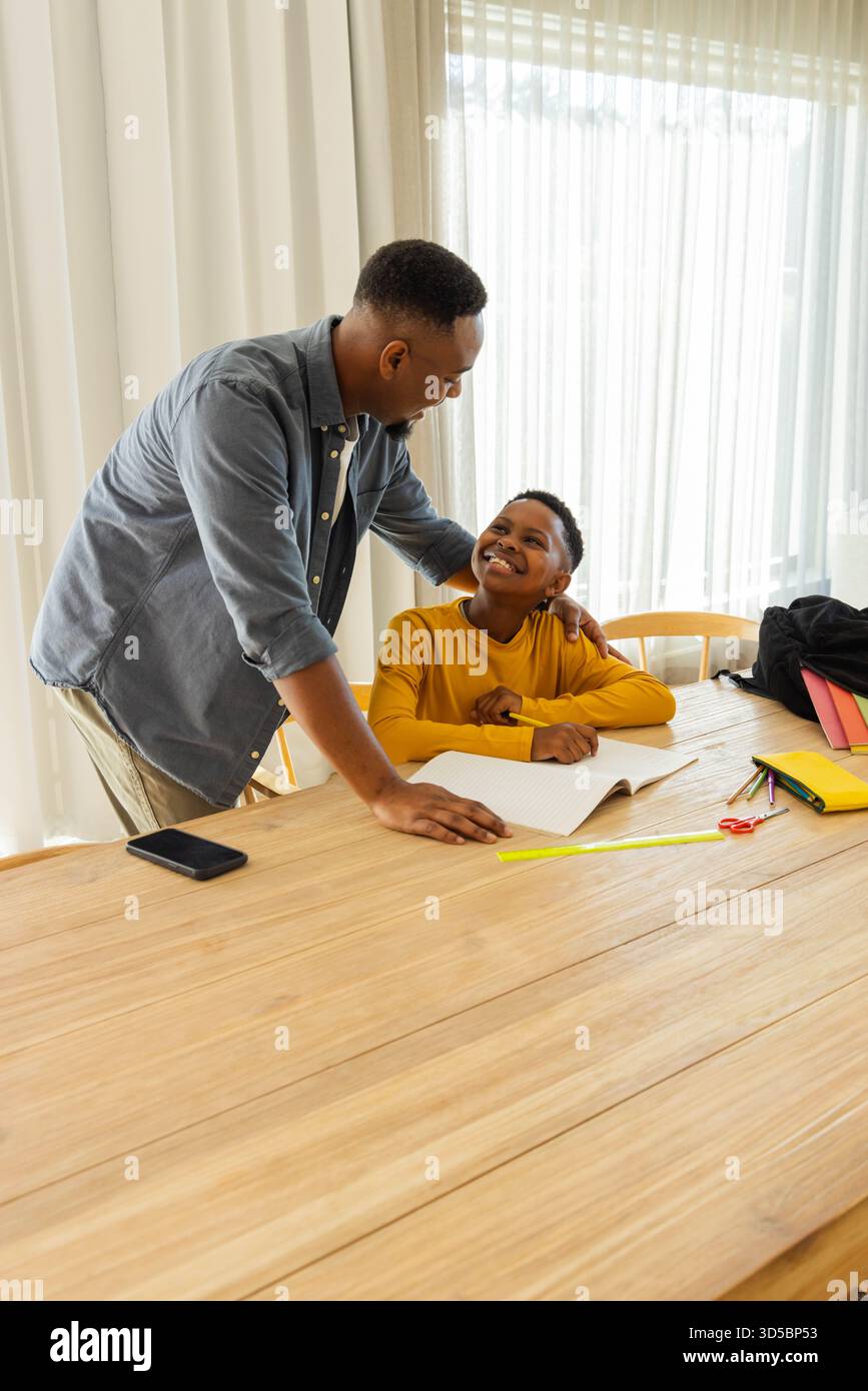 Padre afroamericano che aiuta il figlio con i compiti a tavola, entrambi sorridenti Foto Stock