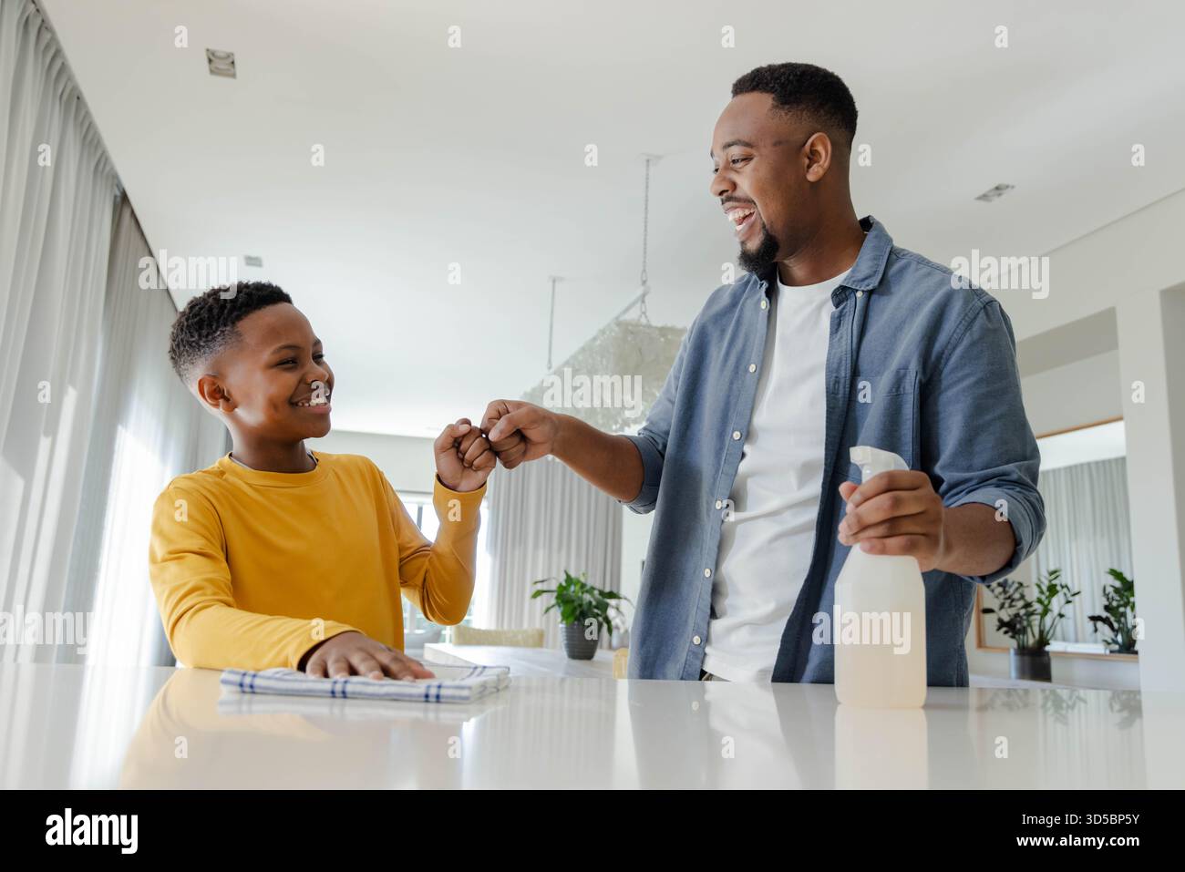 Padre e figlio afroamericani puliscono insieme la cucina, condividendo il pugno e sorridendo Foto Stock