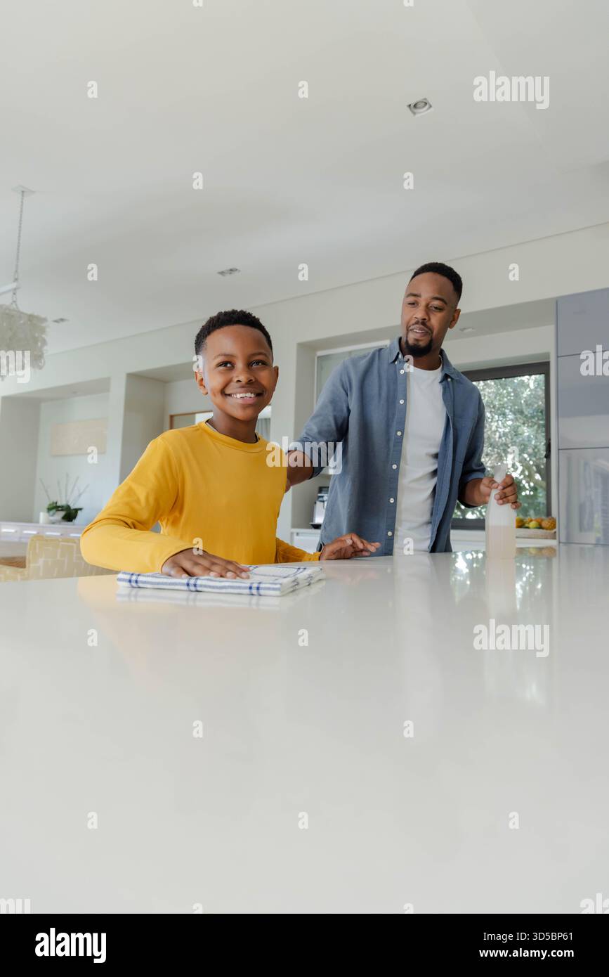 Padre e figlio afroamericani puliscono insieme il bancone della cucina, sorridono e si godono il tempo Foto Stock