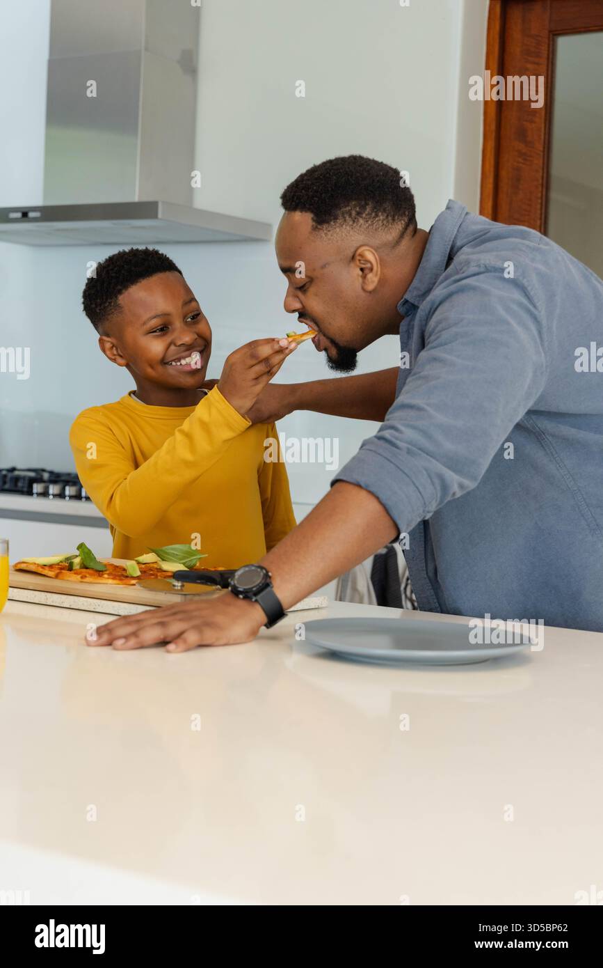 Padre e figlio afroamericani si divertono a cucinare insieme in una cucina moderna, condividendo il pasto Foto Stock